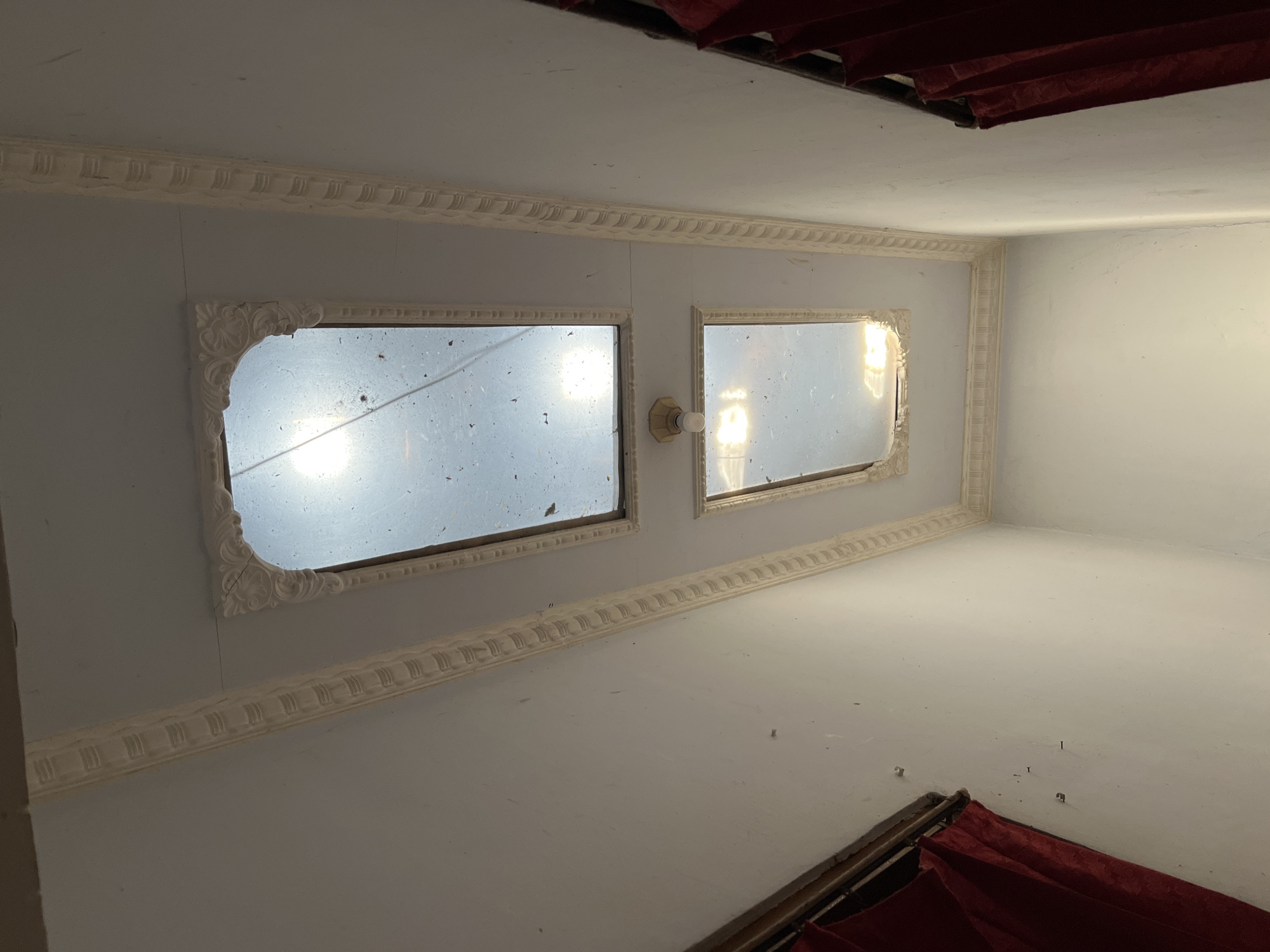 The image shows a ceiling with two skylights and ornate decorative molding around them. The walls are painted white. Red curtains hang on the left and right sides of the image, suggesting windows or alcoves. The ceiling appears dusty, with debris visible on the skylights. A single light fixture is visible between the two skylights. The overall impression is of an interior space, possibly a room or hallway, with natural light entering through the skylights. The scene is consistent with a location in Glagah Kulon, Indonesia, likely an indoor residential or commercial space.