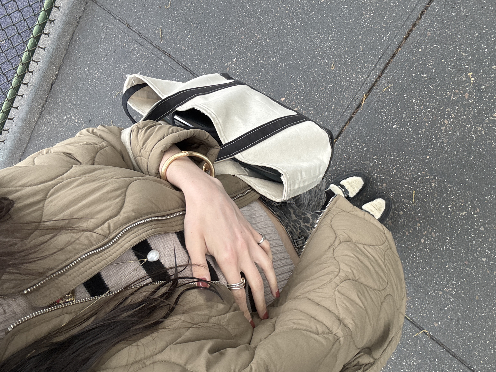 This high-angle, first-person perspective shot looks down at a person outdoors, likely in New York, USA. The subject is wearing a light brown or khaki quilted jacket over a striped sweater, which features black and beige or pinkish stripes. A long necklace with a large white pearl pendant hangs centrally over the sweater.
The person's left arm is visible, adorned with a gold-colored bangle bracelet. Their left hand, positioned centrally, displays multiple silver-colored rings on the index, middle, and ring fingers, with fingernails painted red. Long, dark brown hair cascades over the jacket.
Next to the person, a light-colored canvas tote bag with black straps is visible, containing a dark electronic device, possibly a laptop or tablet. At the bottom right, the person's feet are partially shown, wearing black and cream/white furry slippers or shoes.
The ground beneath is dark gray asphalt, speckled with lighter aggregates and marked by several cracks. A green chain-link fence is visible along the left edge of the frame, suggesting a public outdoor space such as a park or urban street. The attire suggests cool or overcast weather. No text is visible in the image.