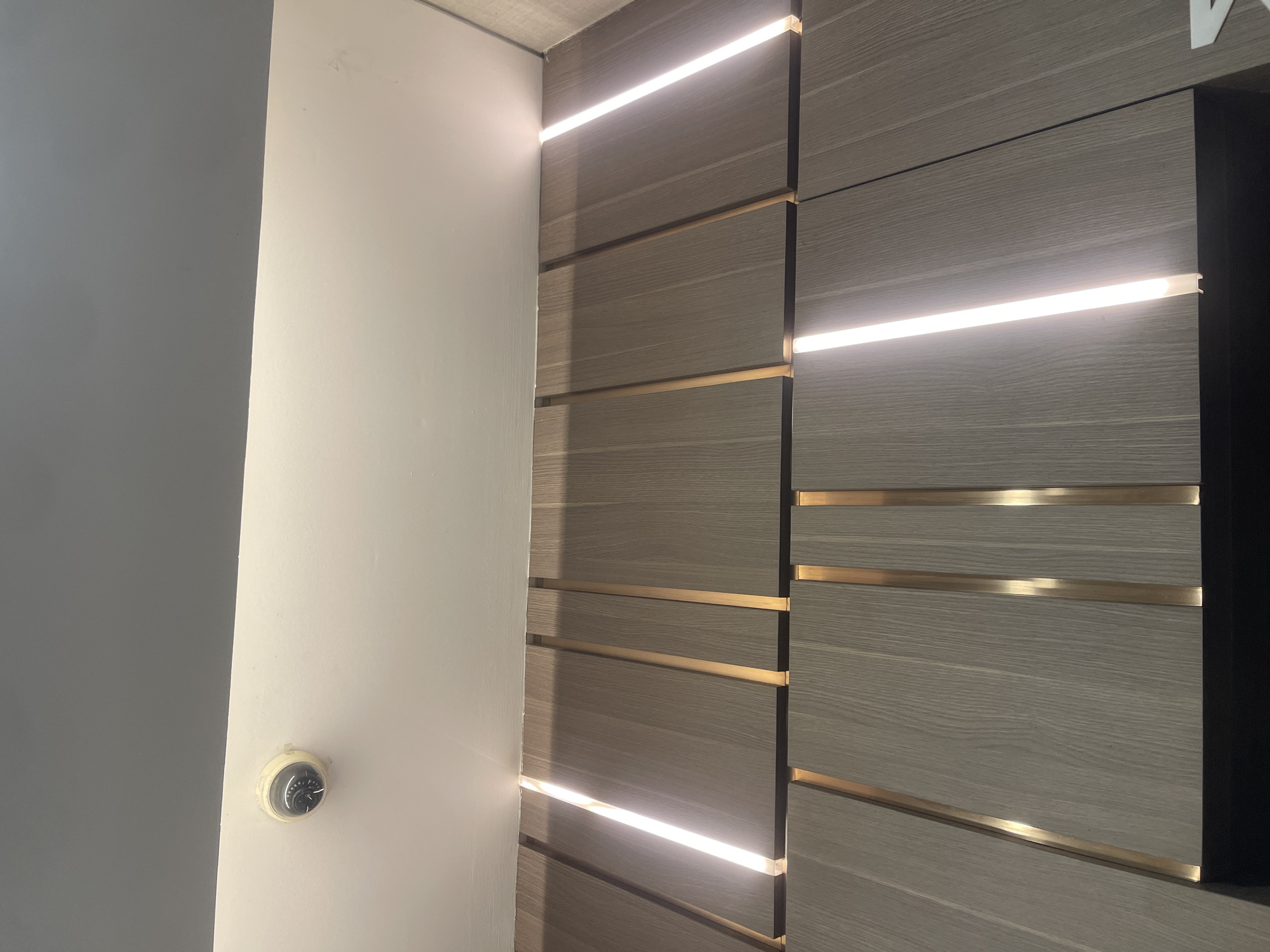 The image shows a close-up, low-angle view of a ceiling and a wall with wooden paneling and integrated lighting. A white dome-shaped security camera is visible mounted on the white ceiling, near the junction with a dark gray ceiling panel. The wall is covered with vertical wooden panels, interspersed with thin, vertical golden strips. Integrated LED strip lights are present in the gaps between some of the wooden panels, casting a bright white light. The bottom of the image reveals a dark surface, possibly the top of a cabinet or furniture. The scene appears to be an interior space, likely a modern or updated building. There are no people visible in the image. This scene is not specific to Kebonagung, Indonesia.