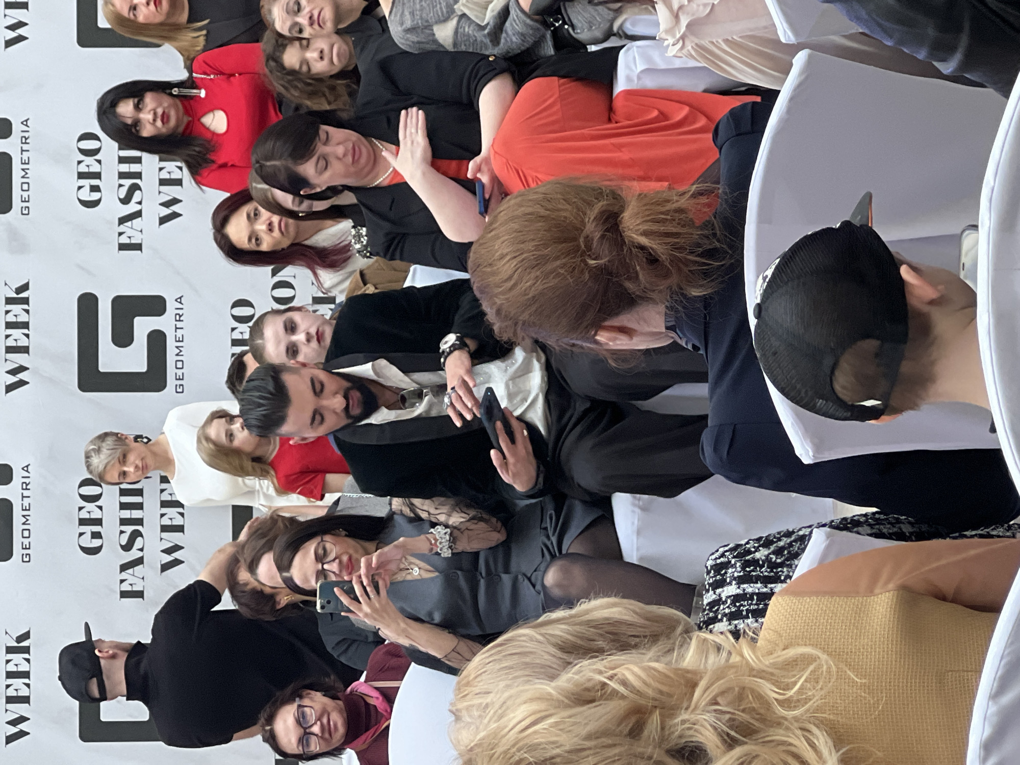 The image captures an indoor scene during a fashion event in Saint Petersburg, Russia. A crowd of seated individuals are attending a "GEO Fashion Week" show, based on the large backdrop behind them. Some attendees are actively using their mobile phones. Men and women of varying ages are present, with diverse clothing styles and expressions. The seating consists of white, cushioned chairs. The setting appears to be in a closed venue, possibly a large hall or auditorium.