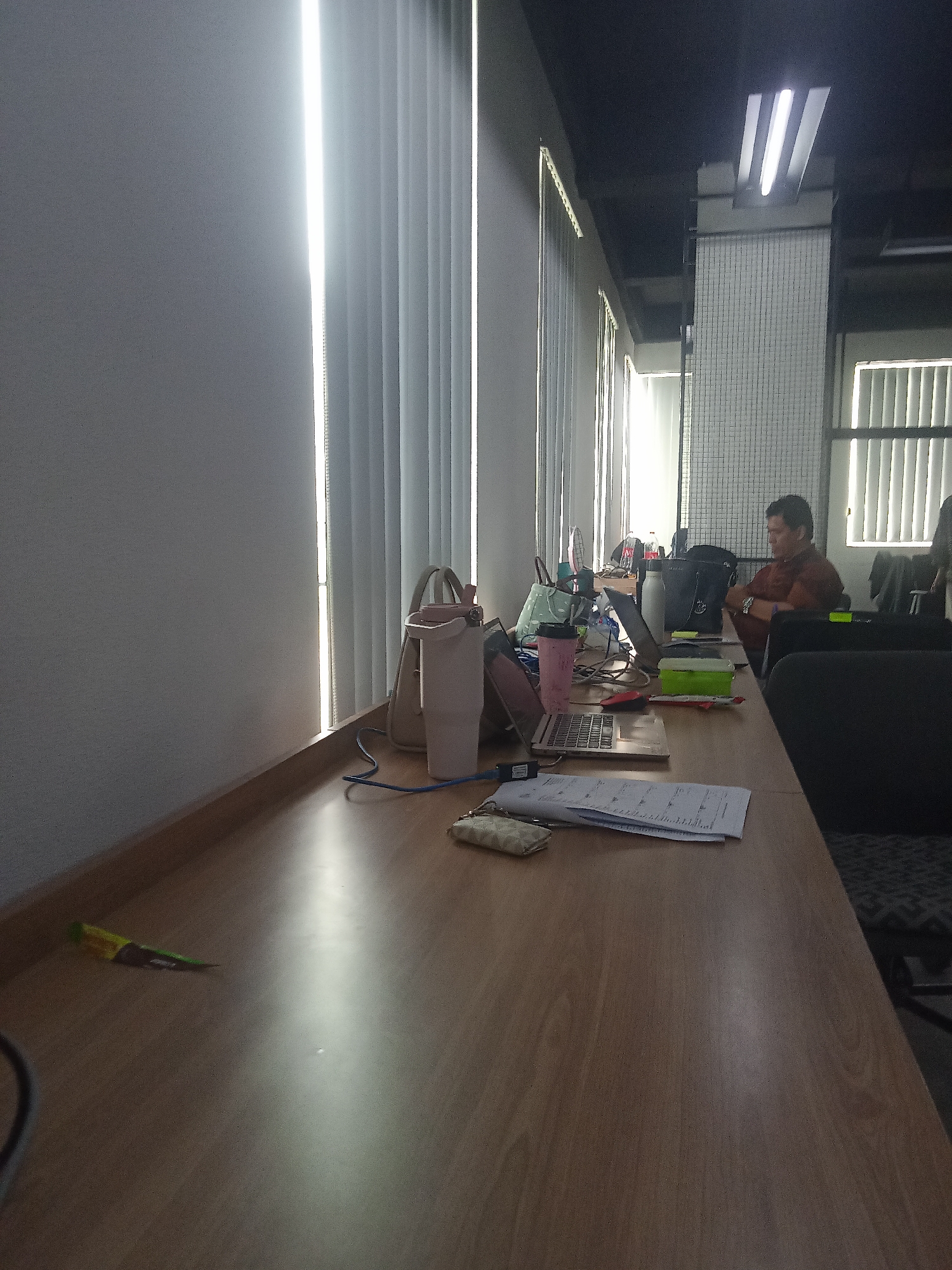 The image shows an indoor office environment, likely in South Tangerang, Indonesia, based on the provided context. A man is seated at a wooden desk, looking towards a laptop. The desk is cluttered with various office supplies, including a laptop, a pink water bottle, a green box, and papers. Sunlight streams in from vertical blinds on the left side of the frame, creating bright vertical lines. The ceiling is dark and features fluorescent lighting. A portion of a dark chair is visible in the foreground. The overall scene suggests a typical workday in a modern office setting.