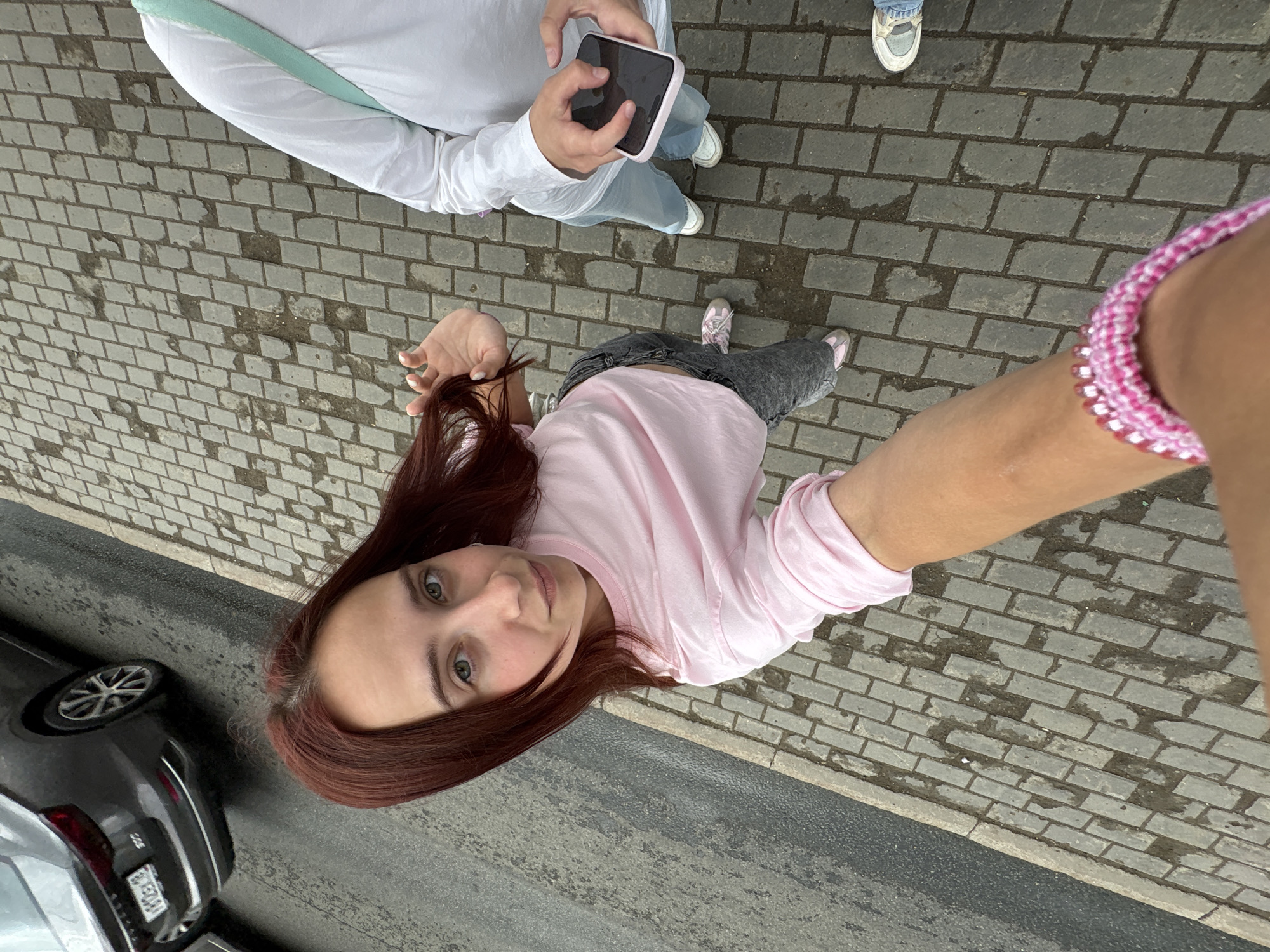 A first-person perspective selfie taken from a high angle shows a young woman with long, reddish-brown hair standing on a grey cobblestone sidewalk. She is wearing a light pink long-sleeved t-shirt and dark grey jeans. Her right hand is raised towards the camera, and she is looking directly at it. To her right, another person, partially visible, is wearing a white long-sleeved shirt and blue jeans. This person is holding a smartphone in their right hand, looking down at it. The background shows a dark grey car parked on the left side of the frame, with its rear end visible, and a wet asphalt road. The image appears to have been taken in an outdoor urban environment, possibly in Izhevsk, Russia, given the style of the car's license plate, which reads "300". The overall scene depicts a casual moment, possibly a brief encounter on a city street.