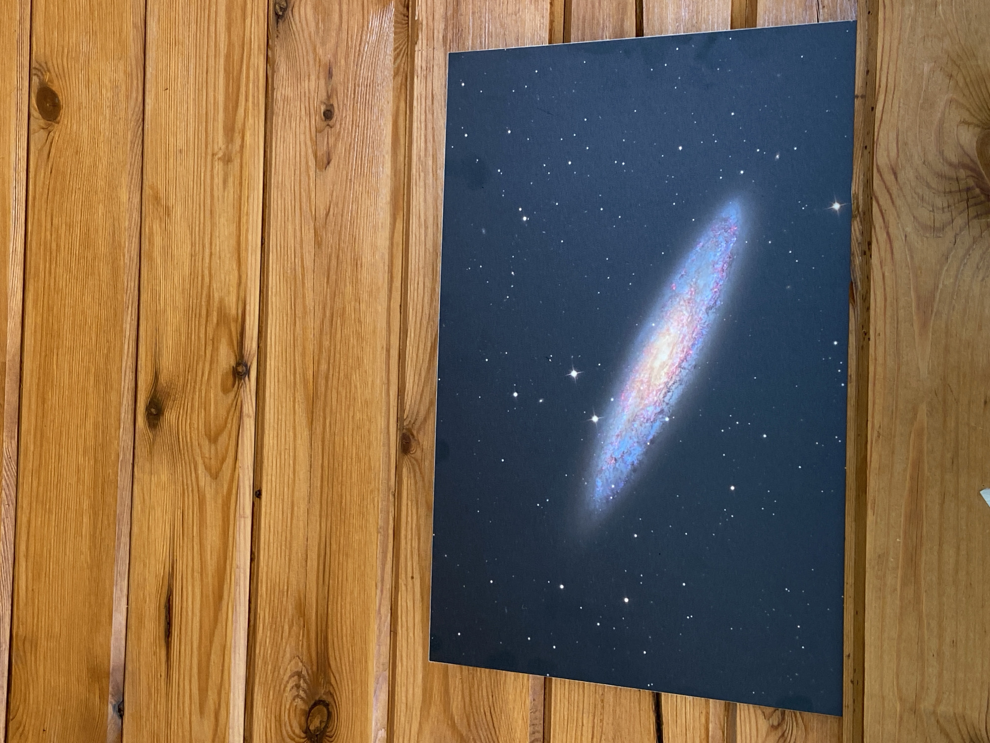 A rectangular print featuring a depiction of the Andromeda galaxy against a dark, star-filled sky rests horizontally on a wooden surface. The surface appears to be made of horizontal wooden planks, possibly part of a wall or shelf, with visible wood grain and knots. The print itself is centered on the wooden surface, taking up a significant portion of the frame. The galaxy is shown as a spiral structure with luminous arms containing vibrant pink and blue hues, surrounded by numerous white dots representing stars. The background of the print is a deep blue-black. The scene is devoid of any people or specific urban environment details.