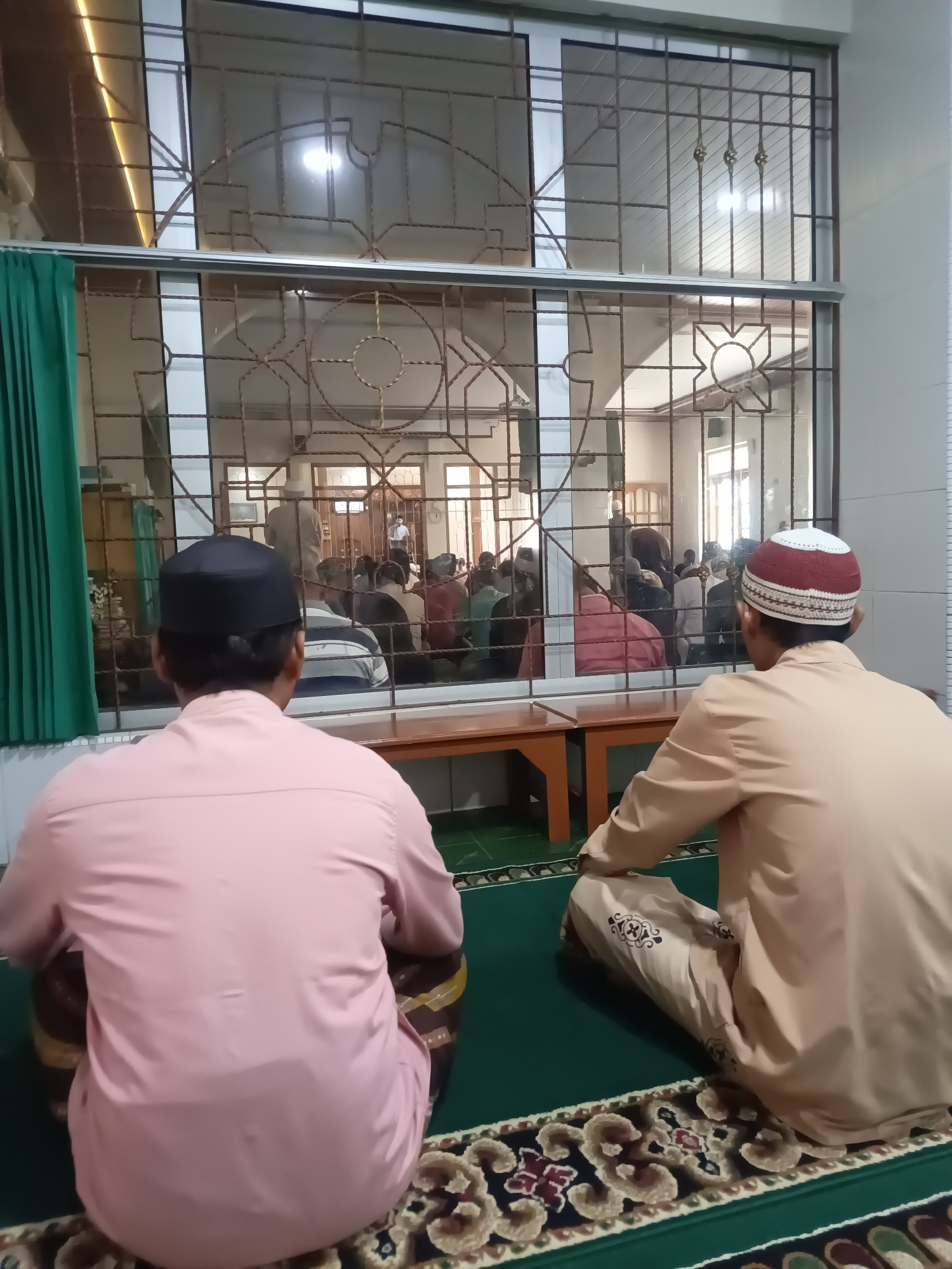 This image depicts a religious gathering taking place in a room at Desa Gunungsimping, Indonesia. Two men, seen from behind, are in the foreground, facing a group of people in the background. The man on the left wears a pink long-sleeved shirt and a black peci (a traditional cap). The man on the right wears a light brown long-sleeved shirt and a white and red peci. Both men are sitting on a green carpet with a decorative border.
Behind them, separated by a decorative metal-framed window or partition, is another room where a congregation is gathered. People are seen sitting on the floor, facing a direction that suggests prayer or a religious sermon. The background room features white walls, a patterned ceiling, and windows with similar decorative metal grilles. A wooden bench or table is visible in the middle ground, positioned beneath the window partition. A green curtain hangs on the left side of the frame, partially obscuring the background on that side. The lighting in the scene appears to be artificial, with a warm glow emanating from overhead fixtures.