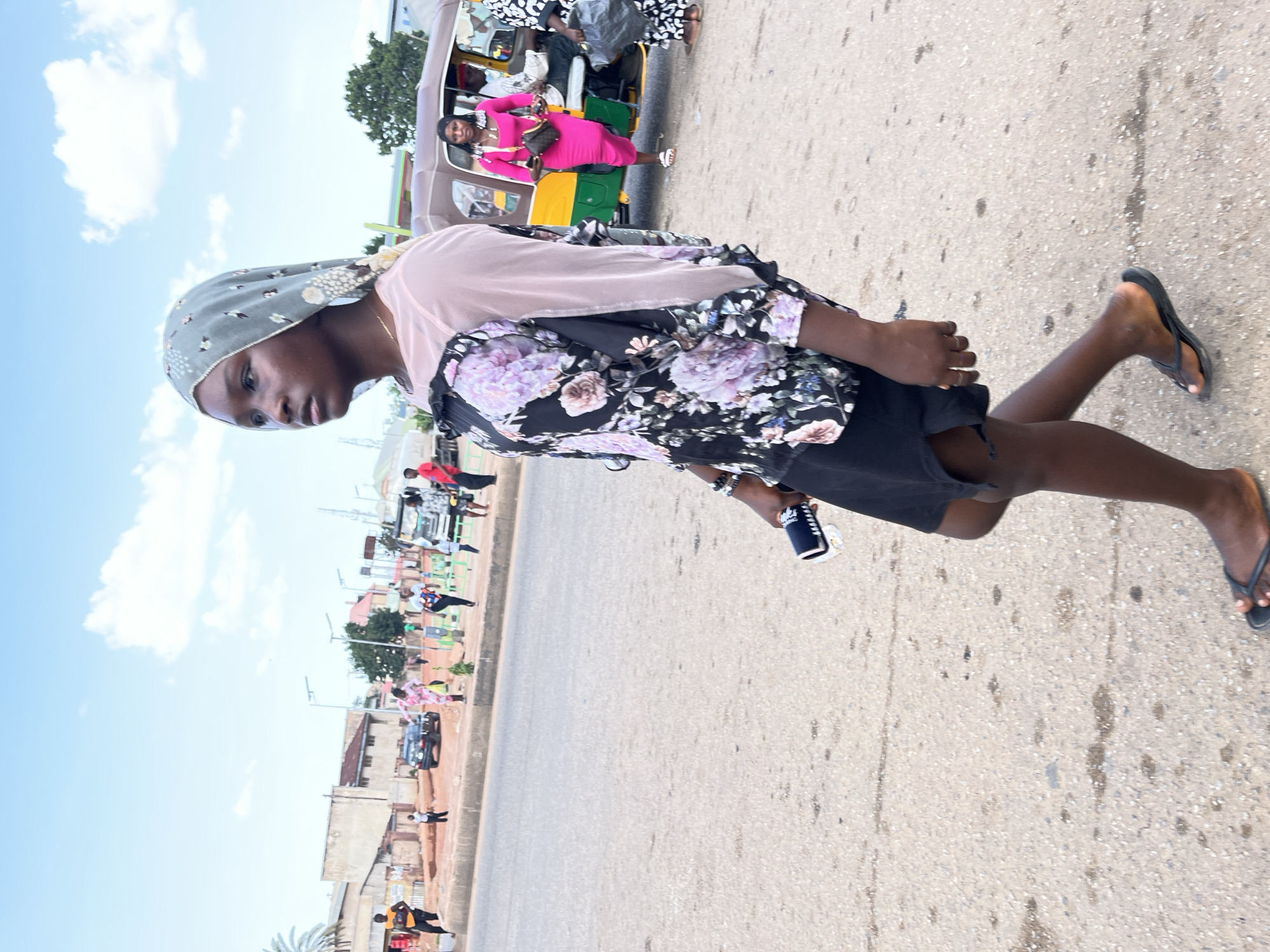 A full-shot, eye-level, outdoor photograph captures a young woman walking on a paved road in Gura Top, Nigeria. She is positioned in the foreground, facing right, with her head turned slightly left to look forward. She wears a light gray hijab with a patterned design, a long-sleeved blouse featuring a light pink sheer upper section and a black lower section adorned with a floral print of purple and white flowers, and a short black skirt. She holds a black object, possibly a phone, in her right hand. Her left arm is bent at the elbow. She is barefoot, wearing black flip-flops on her feet.
In the middle ground, several people are visible near a street. To the right, a woman in a bright pink dress stands near a yellow and brown auto-rickshaw, holding a brown purse. Another person in a black and white patterned outfit stands next to her, carrying a large black bag. The auto-rickshaw appears to be stopped or moving slowly. Behind them, other individuals and vehicles are present, including a white truck. The background consists of buildings and trees under a cloudy sky. The road surface is asphalt with some patches of lighter concrete or gravel.