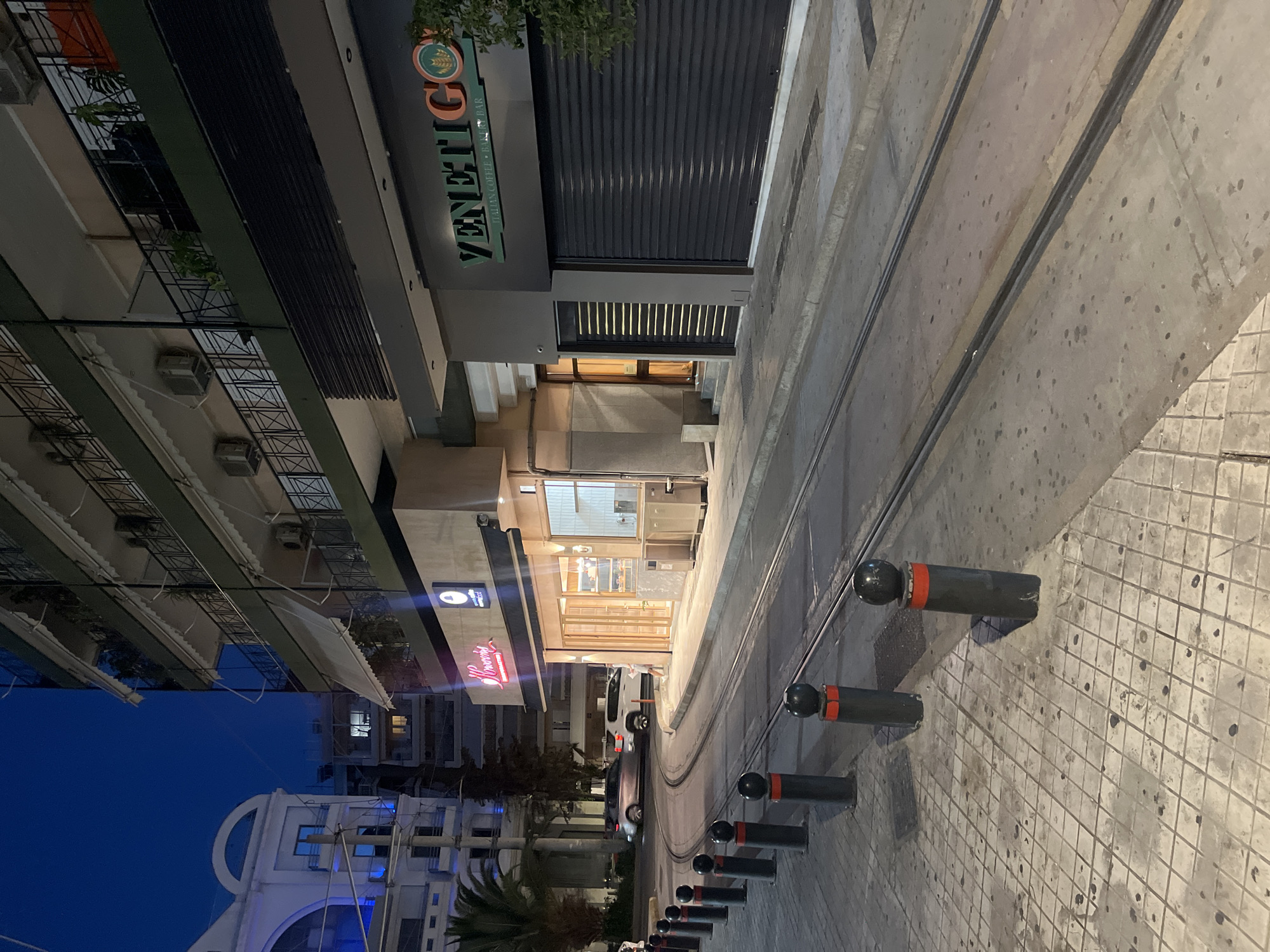 This is a low-angle, eye-level, full exterior shot of a street scene in Athens, Greece, at dusk. The street is paved with large square tiles on the left side, and concrete with tram tracks running down the middle. A line of black bollards with red tops separates the sidewalk from the street. On the right side of the street are two businesses. The first business has a neon sign that reads "Brounis". It has large glass windows and an open door, with a small display of pastries visible inside. The second business has a dark facade with a sign that reads "VENETIGO ITALIAN COFFEE + BAKERY BAR". This storefront has a black metal roll-down security gate. In the background, there are several multi-story buildings with balconies and air conditioning units. Two cars are visible on the street, one silver sedan and one white SUV. The sky is a deep blue, indicating it is twilight. The lighting is artificial, with streetlights and interior lights from the businesses casting illumination on the scene. There is one person visible, a young woman in a white dress, standing on the sidewalk in front of the "Brounis" establishment. The overall impression is a quiet evening street in a commercial district.