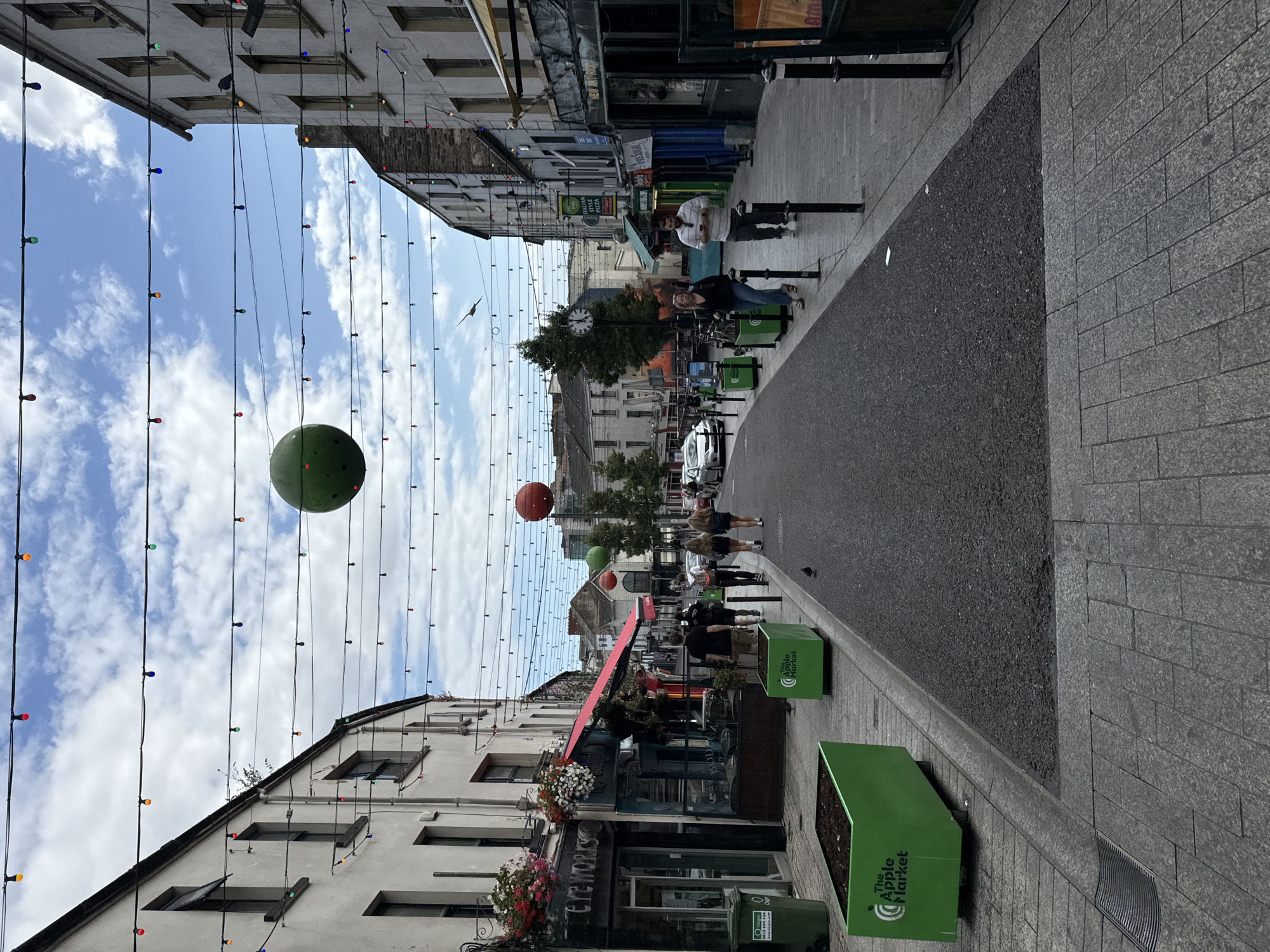 The image depicts a street scene in Waterford, Ireland, likely Georges Street based on the presence of "The Apple Market" planters. The street is lined with multi-story buildings housing various businesses, including "Fireworks" and "Italian Style Pizza". Overhead, strings of colorful lights are strung across the street, with two large spherical decorations, one green and one orange, suspended between them. Several people are walking on the sidewalk, some dressed casually. Cars are visible parked and driving on the street. The sky is cloudy with patches of blue. Green rectangular planters with "The Apple Market" logo are placed along the sidewalk, and a green trash bin is visible on the left side of the image. The street surface is paved with asphalt, and the sidewalks are tiled.