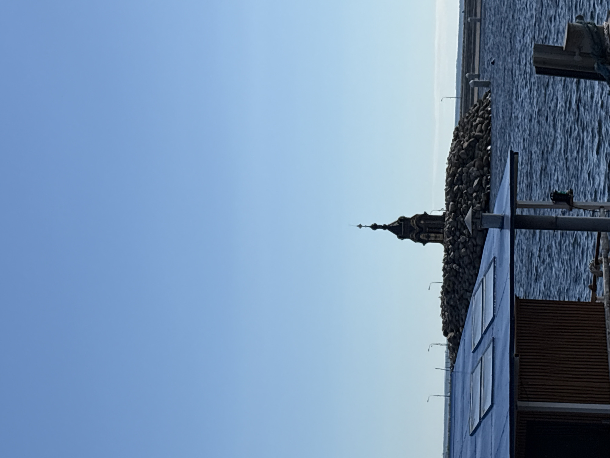 The image depicts a clear, blue sky over a body of water, likely the Gulf of Finland, in Saint Petersburg, Russia. In the foreground, the blue, slanted roof of a building is visible, featuring two skylights. To the left of the roof, a section of a wooden structure is present. In the middle ground, a rocky outcrop forms a small island, topped by a dark, ornate structure resembling a bell tower or a small monument with a cross on top. Several streetlights are sparsely placed on the horizon behind the rocky outcrop, and further out, a breakwater or pier is visible. The water is choppy, with visible ripples. In the lower right corner, a portion of a wooden post and some rope are present, suggesting a dock or pier. The scene is tranquil and depicts a coastal or waterfront area.