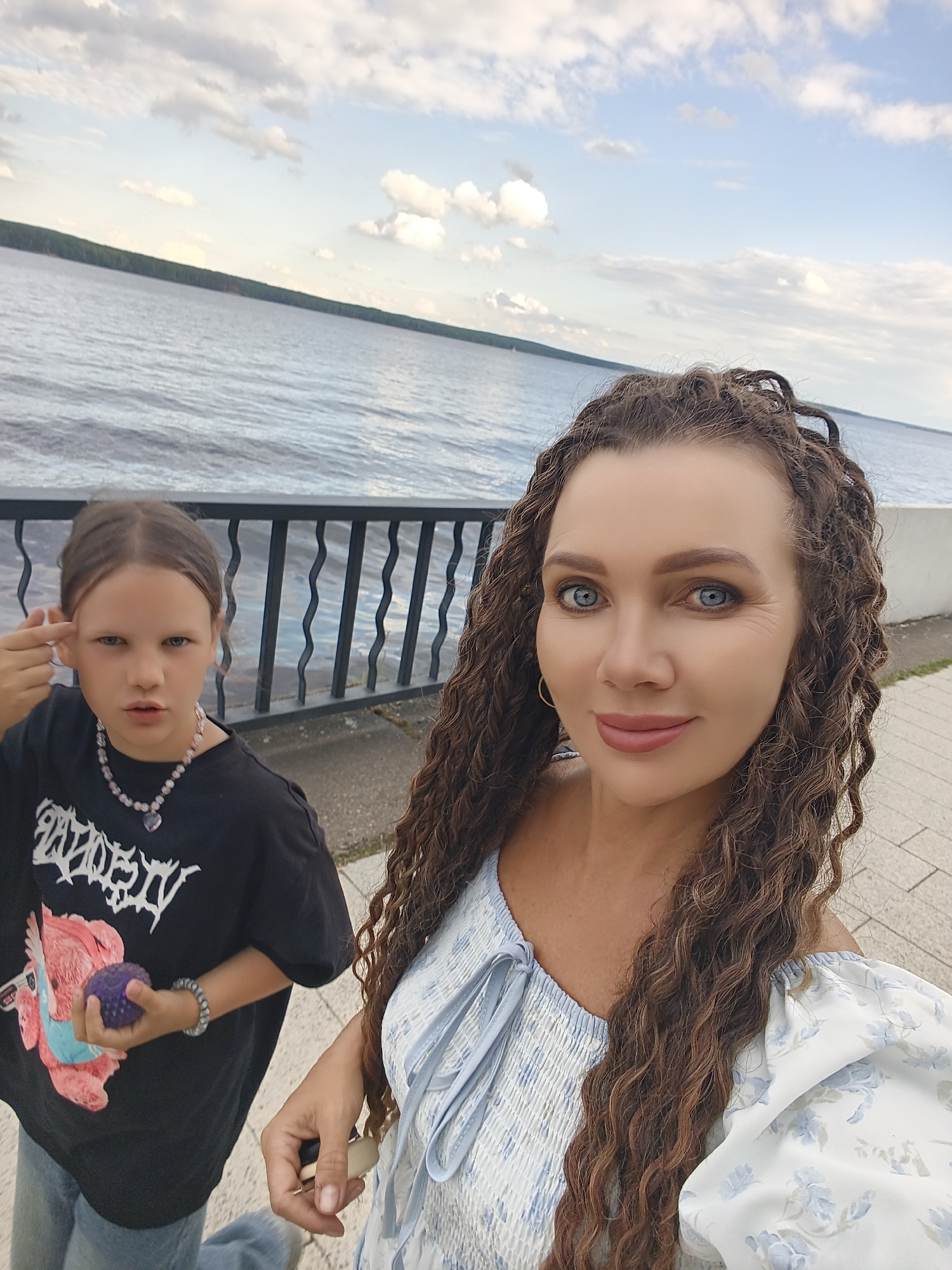 This is a selfie taken outdoors in Chaikovsky, Russia, with a body of water in the background and a cloudy sky above. In the foreground, two individuals are visible. On the right is a woman with long, curly brown hair, blue eyes, and a light blue floral dress. She is smiling slightly and looking at the camera. On the left is a younger person, appearing to be a girl, with brown hair pulled back. She is wearing a black t-shirt with a graphic print and blue jeans. She is holding a small, purple, textured ball in her left hand and has her right hand near her temple, making a gesture. A metal railing and a paved walkway are visible between the two individuals and the water. The water appears calm, with a distant treeline visible across it.