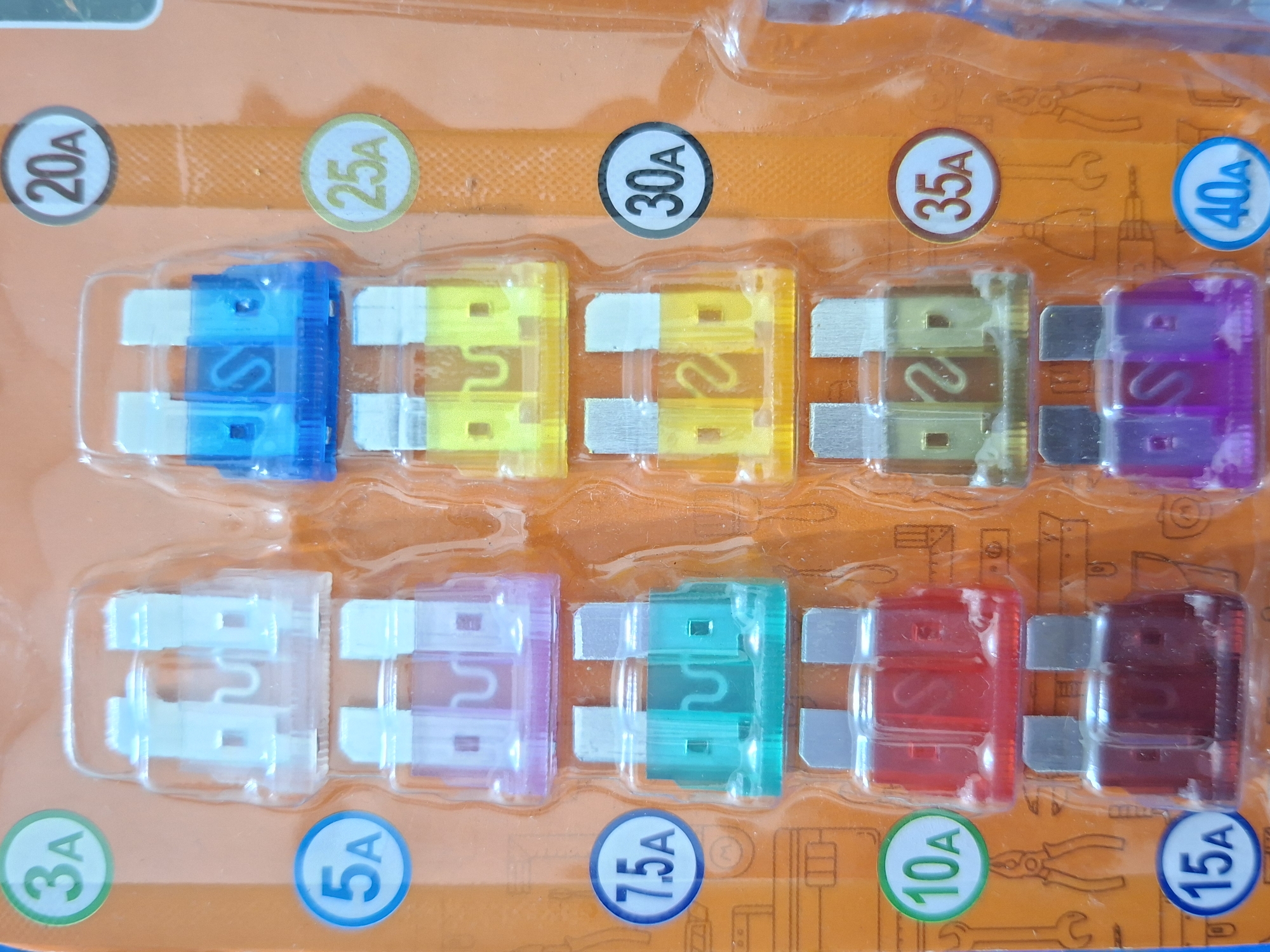 The image shows a variety of automotive fuses arranged in two rows on a predominantly orange packaging. The fuses are of different colors and amperages, ranging from 3A to 40A, with labels clearly visible near each fuse. The packaging is blister-type, with each fuse individually sealed. In the background, under the blister pack, there are illustrated images of tools. There is no information in the image to indicate the location is Lhokseumawe, Indonesia, or any other specific location. The image does not contain any depiction of people.