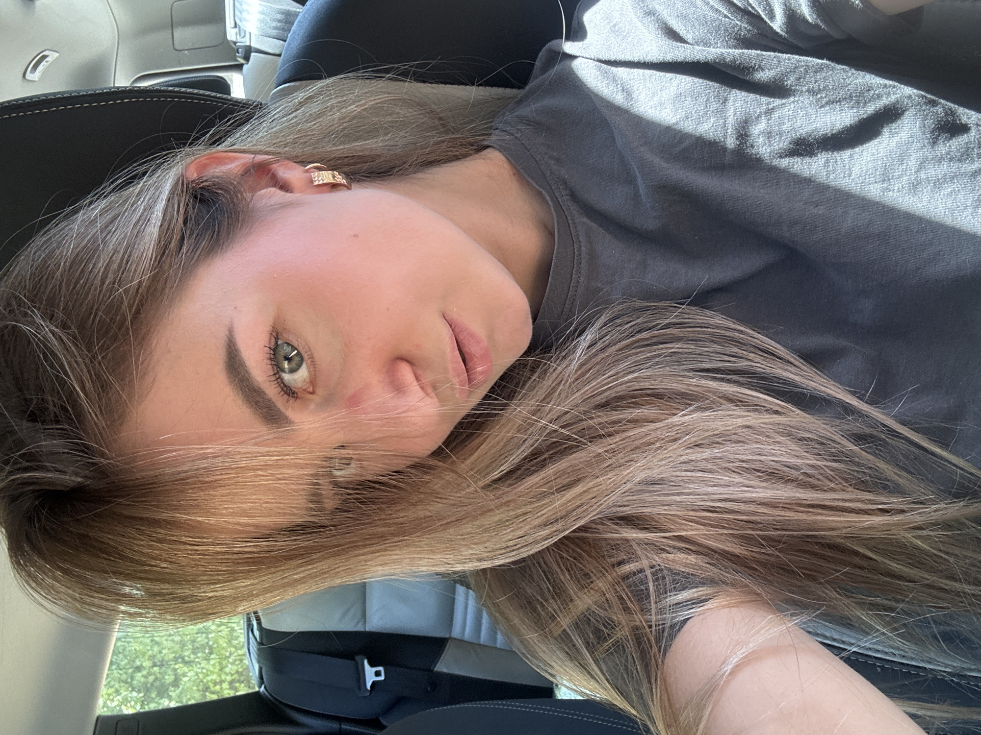 A young woman with blonde hair and green eyes is sitting in the passenger seat of a car. The car interior is visible, with a black leather seat and a seatbelt. Sunlight streams into the car, creating highlights and shadows on the woman's face and dark gray t-shirt. The background shows blurry green foliage, suggesting the car is in motion or parked near trees. The woman is looking towards the camera with a slight smile. The image appears to be a selfie taken inside the vehicle. There is no indication of the location being Saint-Claude, France.