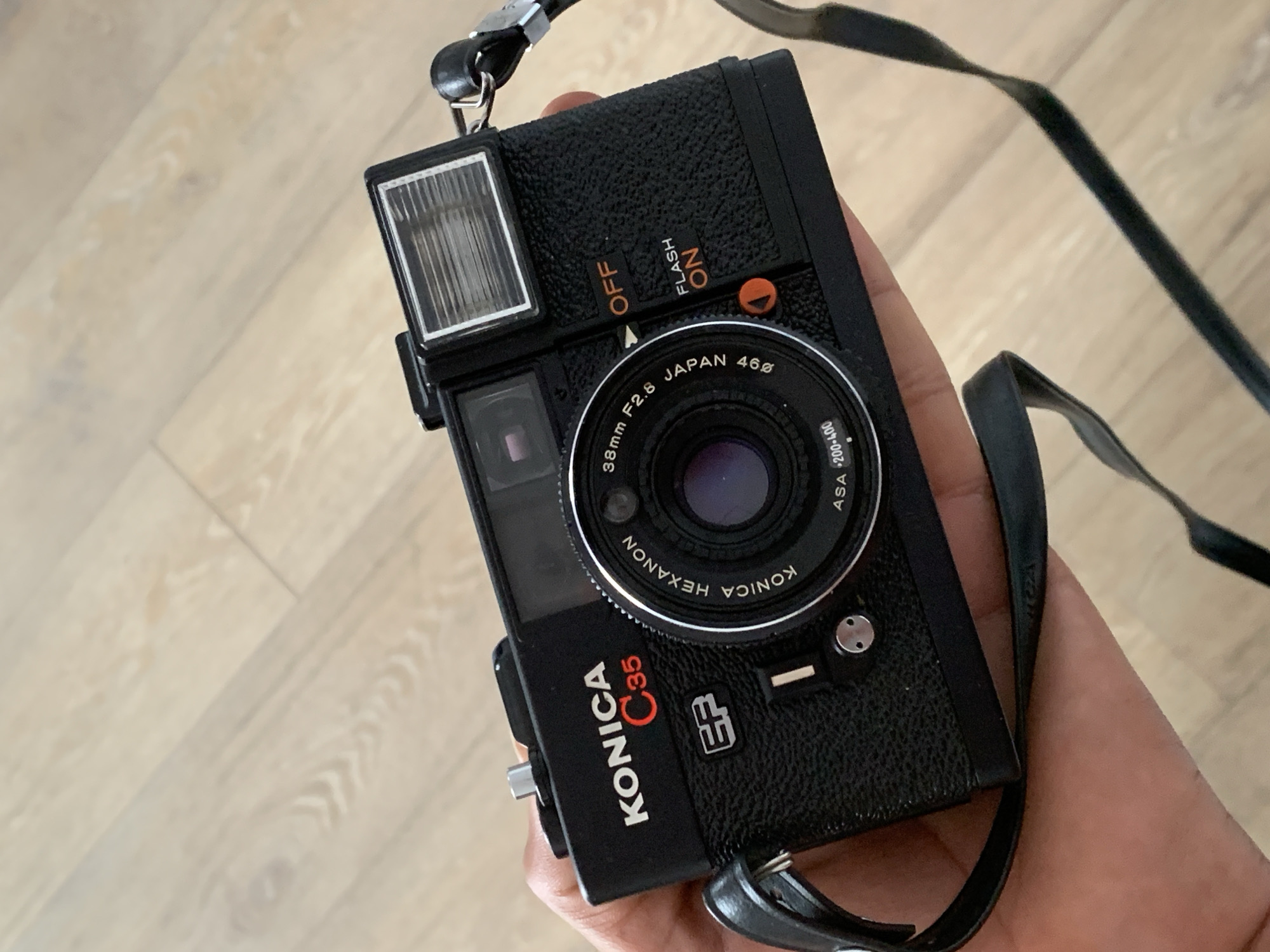 A close-up, slightly angled overhead shot shows a person's hand holding a black Konica C35 camera. The camera has a leather-like texture, with visible branding including "KONICA C35 EF" and "KONICA HEXANON". The lens is visible, along with a flash unit and controls labeled "FLASH ON" and "OFF". The background consists of light-colored wooden floorboards, indicating an indoor setting. There is no identifiable information suggesting the location is Barcelona, Spain.