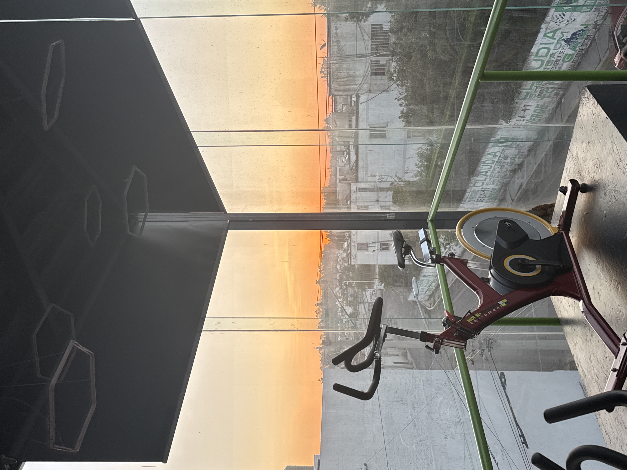 A stationary exercise bicycle sits in a gym with a large window that overlooks the city of Puebla, Mexico. The sky shows a bright sunset. A dark geometric design is visible on the ceiling. An advertisement is visible outside the window on a building.