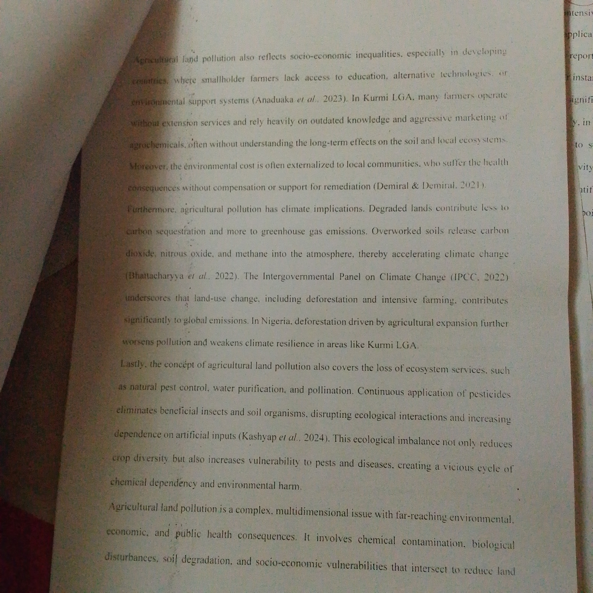 The image shows a close-up of a document with text discussing agricultural land pollution. The text mentions socio-economic inequalities, developing countries, smallholder farmers' lack of access to education and alternative technologies, and environmental support systems in Kurmi LGA, Nigeria. It also details the environmental cost of agricultural pollution, including climate implications, greenhouse gas emissions, and the release of carbon dioxide, nitrous oxide, and methane. The document further explains that agricultural land pollution leads to the loss of ecosystem services like pest control, water purification, and pollination, and discusses the disruptive effects of pesticides on beneficial insects and soil organisms. The text also touches upon the ecological imbalance created, increased vulnerability to pests and diseases, chemical dependency, and environmental harm. Finally, it highlights agricultural land pollution as a complex, multidimensional issue with far-reaching environmental, economic, and public health consequences, including chemical contamination, biological disturbances, soil degradation, and socio-economic vulnerabilities. The background of the image consists of a textured surface, possibly a table or floor, with diagonal lines and shadows, suggesting an indoor setting with artificial lighting. There are no people or specific objects clearly identifiable within the scene other than the document itself. The image is an overhead shot of the document.
