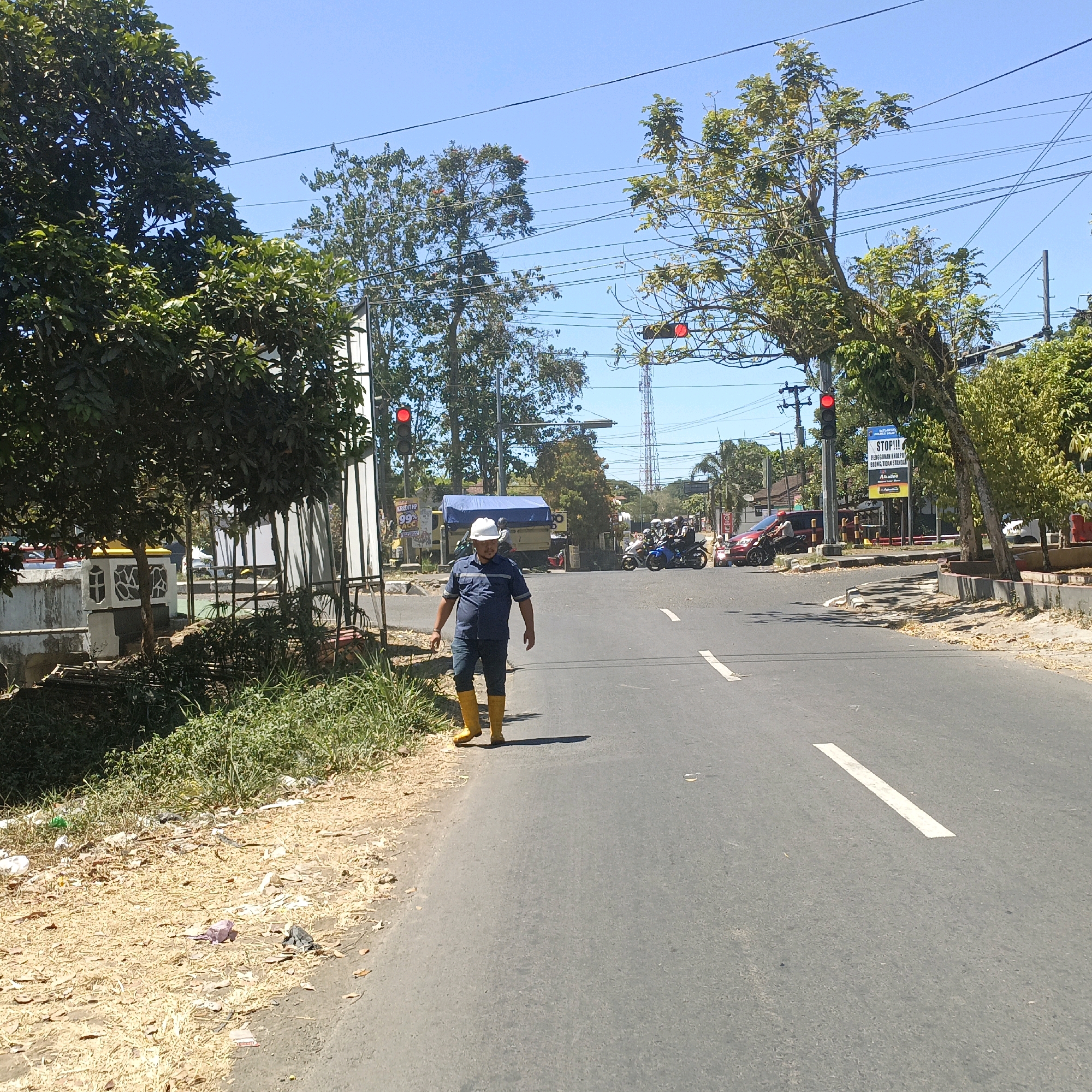 In Salatiga, Indonesia, a man wearing a white hard hat, a dark blue collared shirt, blue pants, and bright yellow rubber boots walks on the right side of an asphalt road. He is walking towards the viewer and slightly to the left. The man is walking on the shoulder of the road where there is dry grass and scattered debris. To his left, the road is empty except for a white line marking the lane. In the background, there is a traffic light that is red, indicating a stop. A yellow truck with a blue tarp is parked on the side of the road to the left of the truck, and a "STOP!!!" sign is visible, along with other advertisements. There are trees on both sides of the road and a clear blue sky overhead. Several motorcycles and a red car are stopped at the intersection.