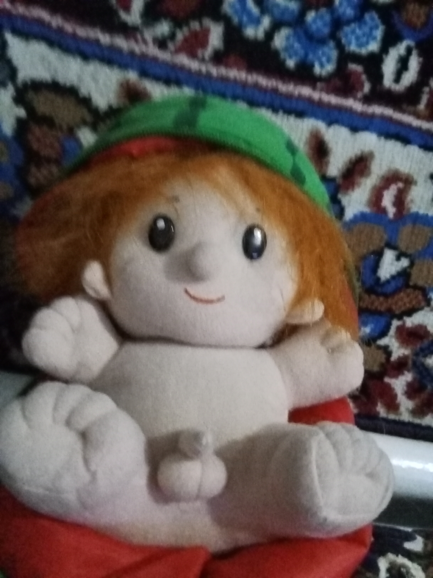 This is a close-up, slightly blurry photograph of a plush toy, likely a doll, sitting on a patterned surface. The toy has orange yarn for hair, a green hat, and a simple embroidered smile. Its eyes are black buttons. The toy is dressed in a red garment that is partially visible. The background appears to be a rug or tapestry with a complex, multicolored pattern featuring geometric shapes and floral motifs in shades of blue, red, white, and brown. The lighting suggests an indoor setting, possibly in natural light. No text is visible in the image. The focus is on the toy, with the background out of focus. The toy's pose and expression are neutral. The location context of Esik, Kazakhstan is not visually apparent from the image itself.