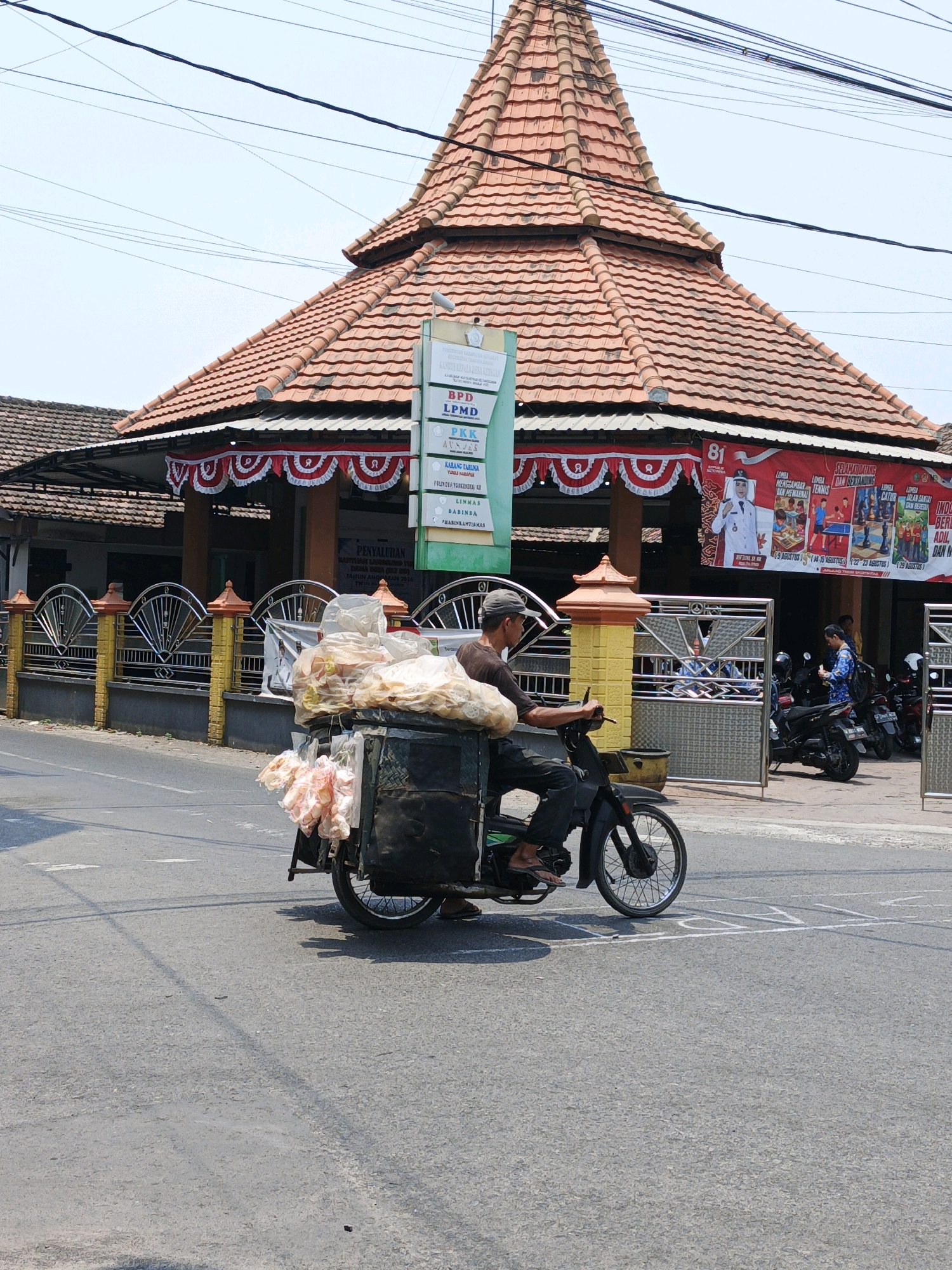 An outdoor, daytime, eye-level shot captures a man riding a motorcycle with a cart in the city of Ketegan, Indonesia. The man, wearing a dark t-shirt and pants, is on a black motorcycle with a cart attached. The cart is loaded with bags of food items, including what appears to be fried snacks wrapped in plastic. In the background, a building with a distinctive orange-tiled, multi-faceted roof stands prominently. This building features a white and green sign with text in Indonesian, and a large red banner is displayed across its facade, decorated with white and red pennants. The banner includes an image of a person in a white uniform and text related to an anniversary or event, with smaller inset images depicting sports activities. The area appears to be a street scene, with a paved road in the foreground and several motorcycles parked in front of the building. A yellow and black metal fence with decorative elements runs along the side of the building. The sky is clear and blue, with some power lines visible.