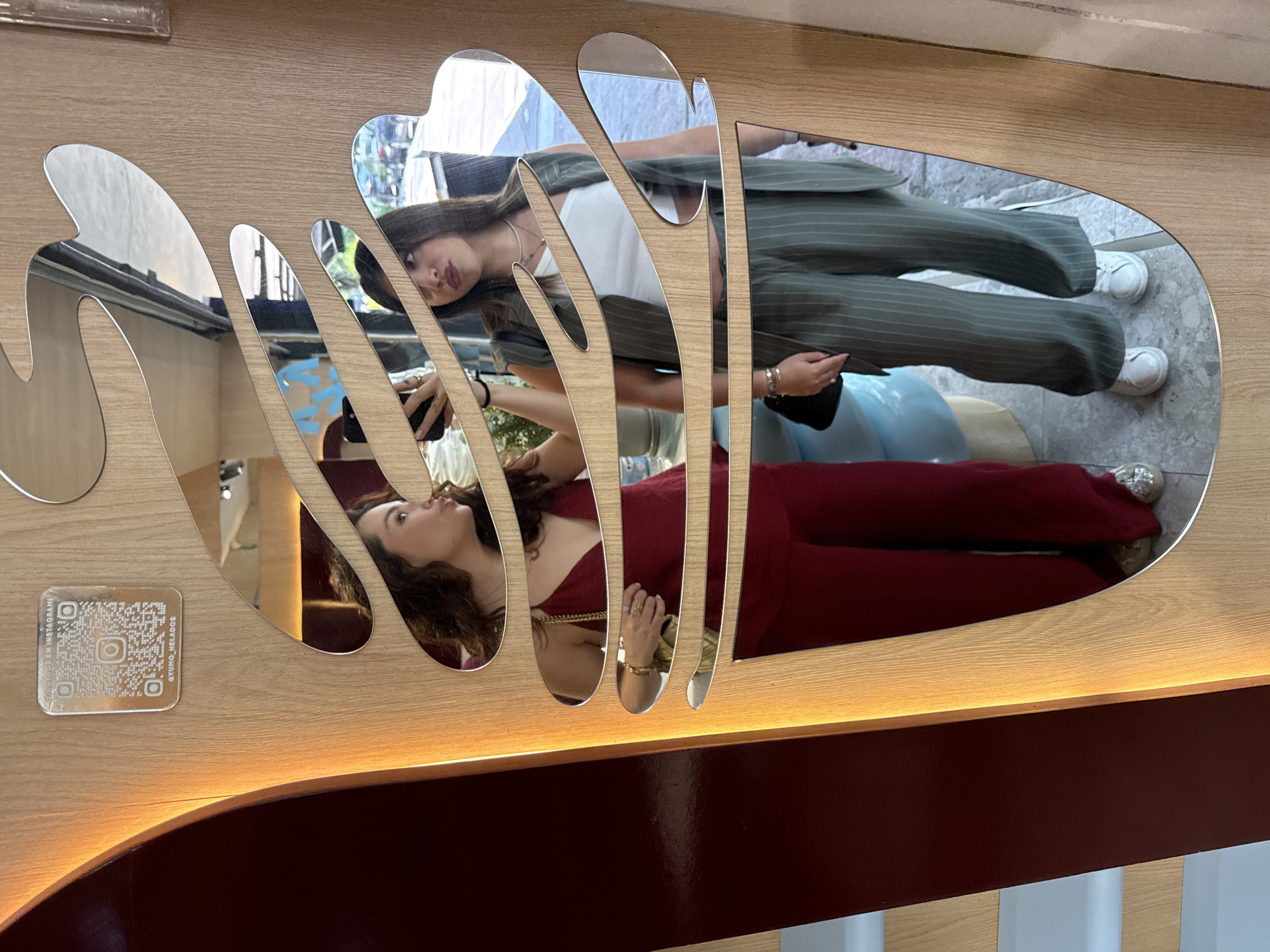 The image captures a reflection of two women in a uniquely shaped mirror on a light brown wooden surface. A QR code and text, presumably an Instagram handle, are affixed to the wood near the mirror's edge. The reflection shows the first woman dressed in a red jumpsuit while the second is in a white top and striped green suit. They are standing in front of a bright, neutral backdrop, possibly a public space. Based on the presence of the QR code labeled with "@yumo_helados," it's likely the photograph was taken in Mexico City, Mexico, at a business called Yumo Helados.