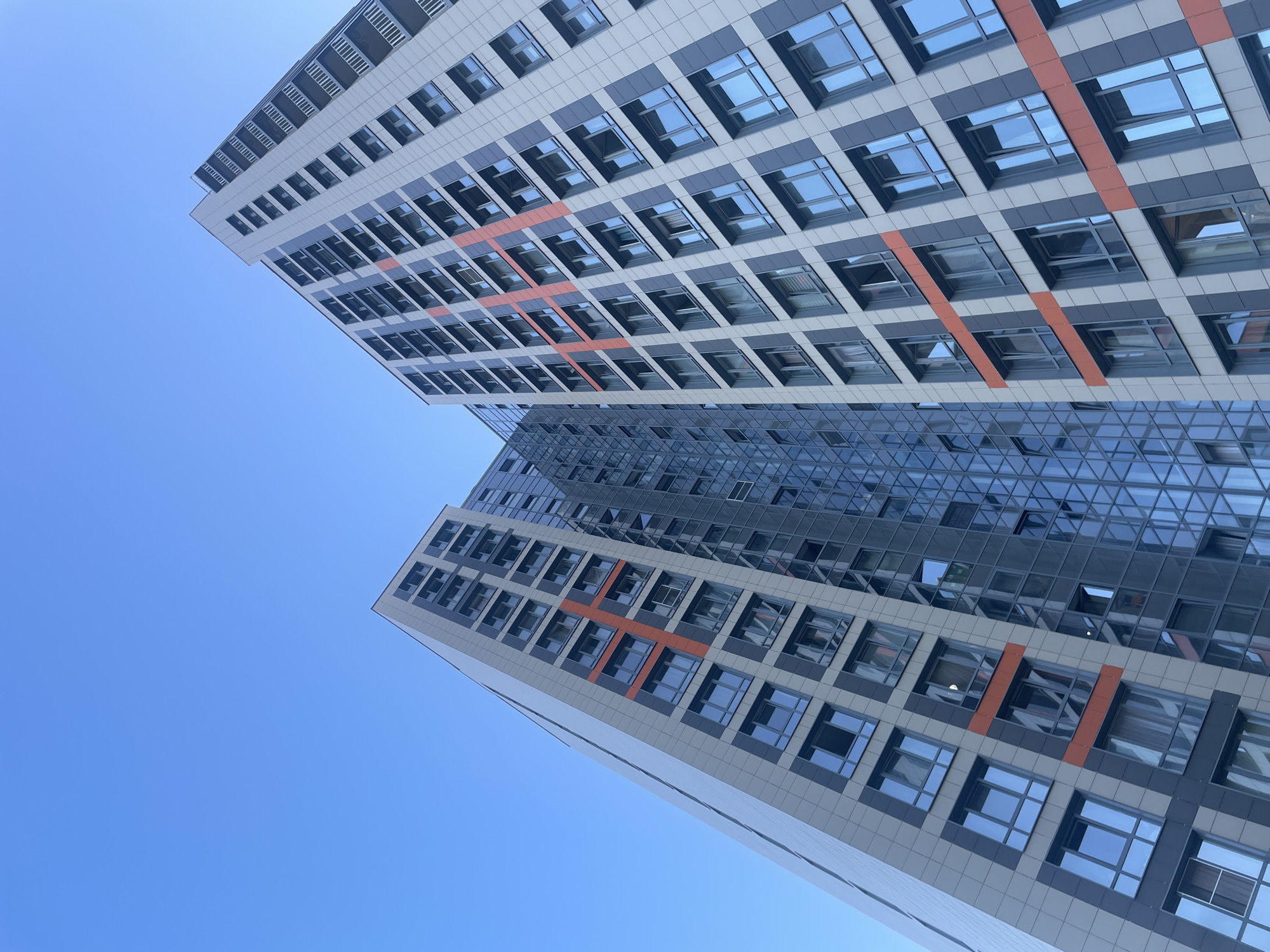The image shows the exterior of two modern high-rise buildings with a clear blue sky as the background. The buildings are predominantly grey and white, with dark grey and orange accents. Both structures feature numerous windows, with some reflecting the sky and others revealing interior lights or blinds. One building has a section with a glass facade, creating a mirrored effect of the adjacent building and the sky. The perspective is from a low angle, looking upwards, emphasizing the height of the buildings. There are no people or vehicles visible in the frame. The architecture suggests a contemporary urban environment, likely a residential or commercial complex.