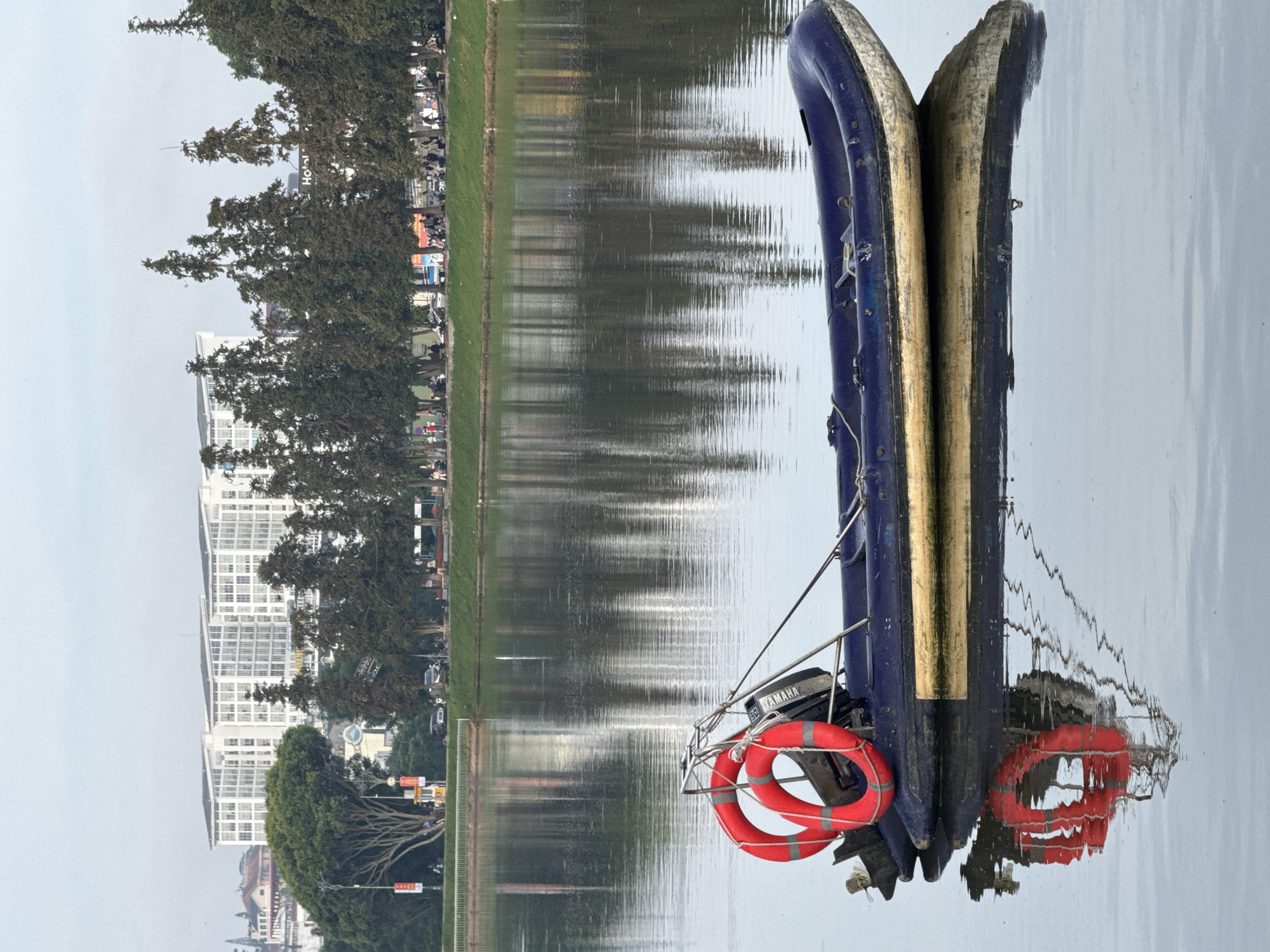 Two inflatable boats are moored on a body of water. The boat on the left is a dark blue inflatable dinghy with a golden stripe along the top. It has a small black outboard motor attached to the back. A red life preserver with white stripes is secured to the side of the boat, hanging near the motor. The boat on the right is partially obscured, but appears to be a similar inflatable boat. The boats are floating on calm water, reflecting the overcast sky.
In the background, a grassy embankment lines the water's edge. Beyond the embankment, trees with dark green foliage are visible, with a large, modern white building with many windows rising behind them. Further in the distance, more trees and buildings can be seen. There are some signs and people visible along the embankment, suggesting it's a public area. The overall scene is one of stillness and quiet. The lighting suggests it is daytime, possibly late afternoon or early morning, given the soft, diffused light and lack of strong shadows. The calm water and overcast sky create a muted and serene atmosphere. The presence of the life preserver suggests recreational use of the boats, possibly for fishing or leisure on the water. The text "YAMAHA" is visible on the outboard motor.