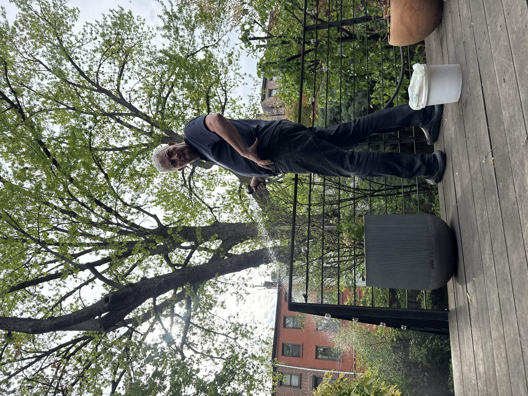 The image depicts a man standing on a wooden deck. He is wearing a dark blue shirt and dark pants, and he has short grey hair. He is holding a watering hose, with water visible in the air. A large tree with green leaves is visible overhead. A black metal railing with spherical ornaments runs along the edge of the deck. Behind the railing, a red brick building with windows is visible. A white bucket with a plastic bag inside it sits on the deck near the man's feet. The scene appears to be a residential rooftop in an urban setting, most likely the city of New York.
