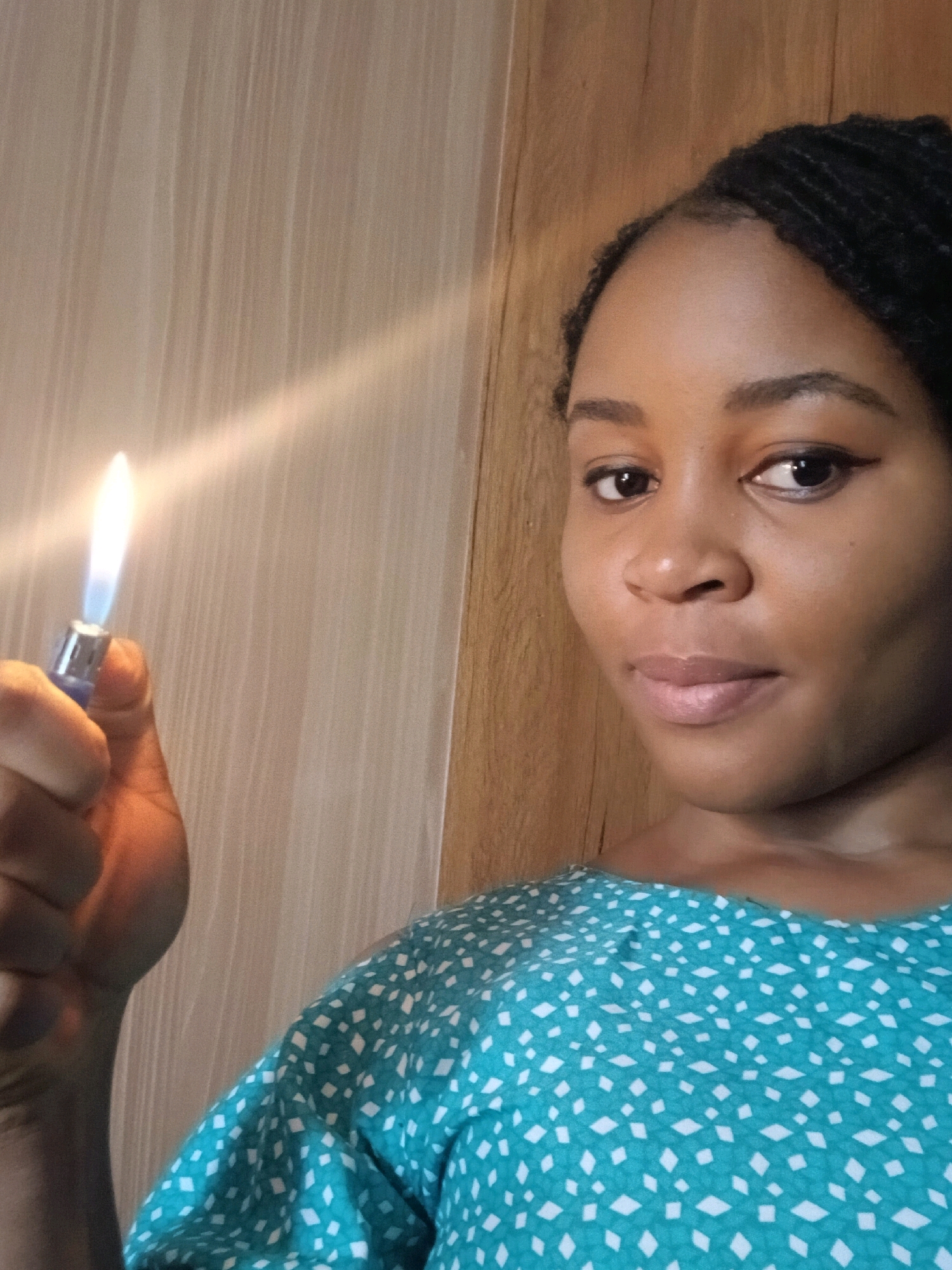 A medium shot shows a Black woman holding a lit blue lighter up to her left. The lighter's flame is bright white and yellow, with a blue base. The woman has braided black hair and is wearing a turquoise top with a white diamond pattern. She is looking at the camera with a neutral expression. The background appears to be a wall with vertical wooden panels on the right and a lighter colored, possibly textured surface on the left. The image is taken indoors, with artificial lighting.
