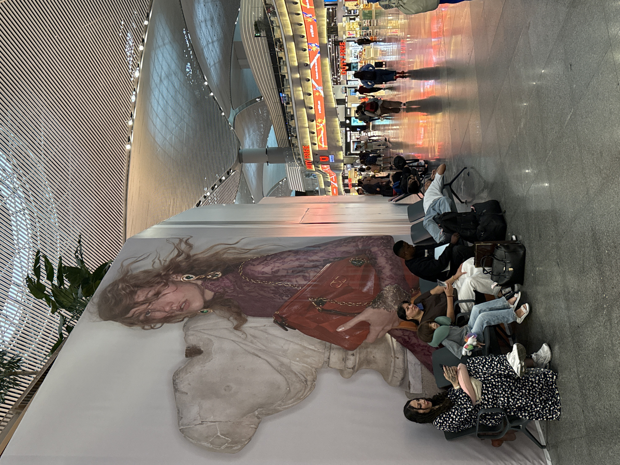 The image depicts a scene within a modern airport terminal, likely Istanbul Airport (IST) in Arnavutköy, Turkey, given the architectural style and branding visible. The terminal features a high, slatted ceiling with integrated lighting, creating a spacious and contemporary atmosphere. A large, impactful mural of a woman holding a red handbag is prominently displayed on the left side of the frame, juxtaposed with a classical architectural fragment.
In the foreground, several individuals are seated on dark-colored waiting area benches. On the far left, a woman in a black and white floral dress is looking at her phone. Next to her, a child with a neck pillow sits on the bench, holding a plush toy. An adult woman sits beside the child, and further down the row, a man in a dark sweatshirt is seated. Another person appears to be resting or sleeping on a bench further down the row, with luggage beside them.
The background reveals a bustling airport environment with shops and signage. "DUTY FREE" signs are clearly visible, indicating retail outlets. People are walking through the terminal, some appearing to be travelers with luggage. The floor is polished and reflective, showing the ambient lighting of the terminal. The overall impression is one of a well-lit, modern, and active transportation hub.