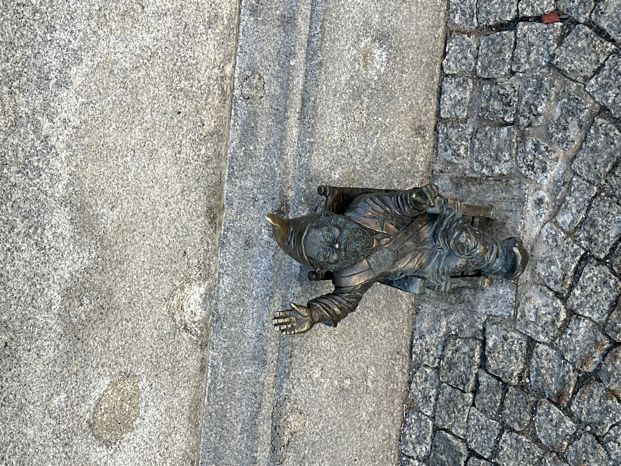 An overhead shot captures a bronze gnome statue situated on a textured ground surface in Wrocław, Poland. The image is taken from a slightly angled perspective, looking down upon the dwarf.
The main subject is a small, detailed bronze gnome figure. It wears a pointed hat, has a prominent beard, and appears to be clad in typical gnome attire, including a tunic and boots. The statue shows some signs of patina, indicating its metallic composition and exposure to the elements. The gnome's right arm (from its own perspective) is bent, holding a small, cylindrical object or tool against its side. Its left arm is outstretched forward with an open palm, as if gesturing or reaching for something on the ground. The gnome's facial expression appears focused and perhaps slightly contemplative or determined.
The setting consists of two distinct ground textures: to the left, a large expanse of lighter, finely-grained grey stone or concrete, and to the right, a section of darker, rougher cobblestone paving. A clear, straight boundary separates these two surfaces, with the gnome positioned directly on this dividing line, slightly encroaching on the cobblestones. The lighting suggests a bright daytime, with subtle shadows and highlights visible on the statue and ground, though no specific weather conditions are discernible. There is no visible text in the image. The presence of numerous gnome statues is a well-known characteristic of Wrocław.