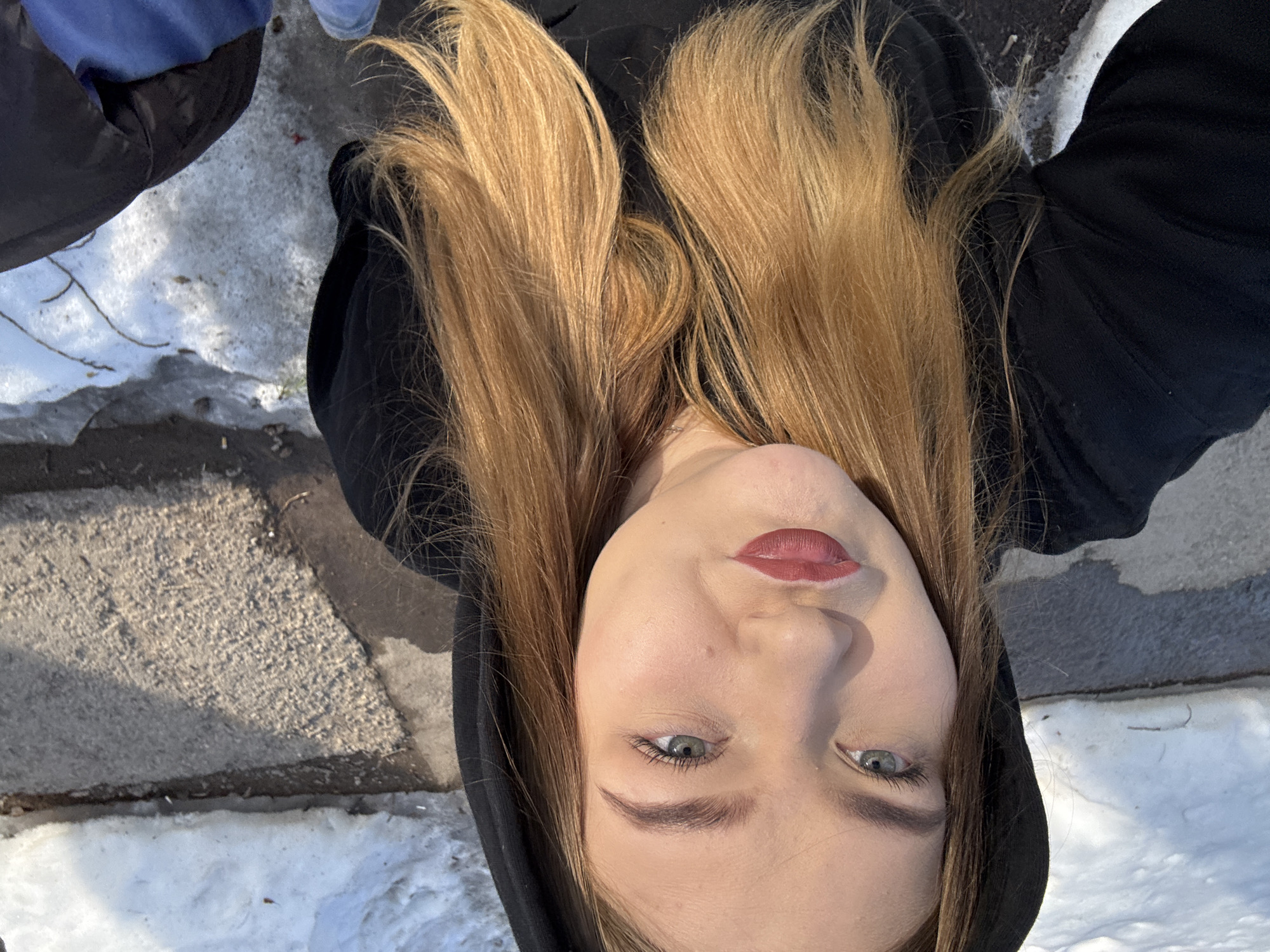 A woman with long blonde hair, wearing a black hooded sweatshirt, is positioned in an outdoor setting. She is looking up at the camera, which is positioned above her. Her face is clearly visible, and she has fair skin, light eyes, and wears reddish-pink lipstick.
The background includes snow and a concrete surface. Fragments of an additional figure (partial blue and black jacket) are visible in the periphery of the frame.
The location is suggested to be in the city of Chaikovsky, Russia.