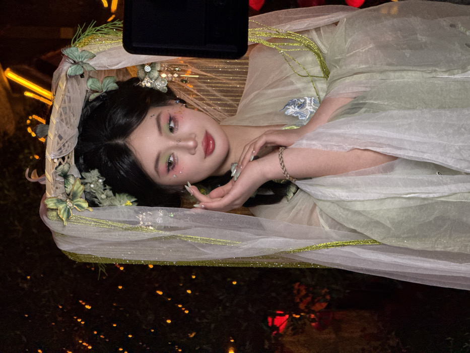 An East Asian female subject is captured in a posed, illuminated portrait within Yuzhong District, China, under low-light conditions. She is dressed in an elaborate traditional Chinese-style costume, predominantly in shades of light green and white. This attire includes a sheer, glitter-infused veil with green linear embellishments draped around her, and a woven headpiece adorned with light green fabric butterflies, artificial flowers, and foliage. Her dark hair is styled in an intricate updo, further decorated with additional ornate elements, including dangling white and silver ornaments on the viewer's left side of her head. Her face features detailed makeup with pink and green eyeshadow, glitter on her cheeks, defined eyebrows, and reddish-glossy lips. Her hands, displaying manicured nails, are held delicately in front of her chest; a braided bracelet is visible on her left wrist. The background is dark and out of focus, showcasing numerous warm orange and red bokeh lights, suggesting a festive or well-lit urban environment with indistinct foliage or structures. A dark, rectangular object, possibly photographic equipment, partially obscures the right edge of the frame. The subject's gaze is directed off-camera to her right with a serene expression.