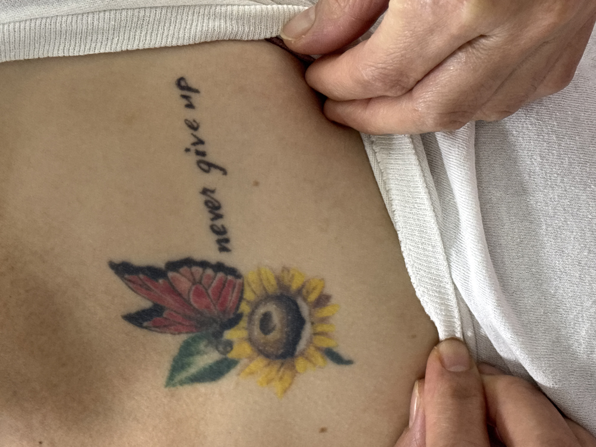 A close-up shot focuses on a tattoo on a person's chest. The tattoo features a butterfly with red and black wings resting on a yellow sunflower with green leaves. To the right of the tattoo, the phrase "never give up" is inscribed in black, cursive lettering. The skin appears slightly tanned. A portion of a white garment, likely a t-shirt, is visible, framing the lower part of the image. Two hands are present in the foreground, pulling the fabric of the garment away from the chest, revealing the tattoo. The background is indistinct. No specific location in Dubai, United Arab Emirates, can be determined from the visual information provided.