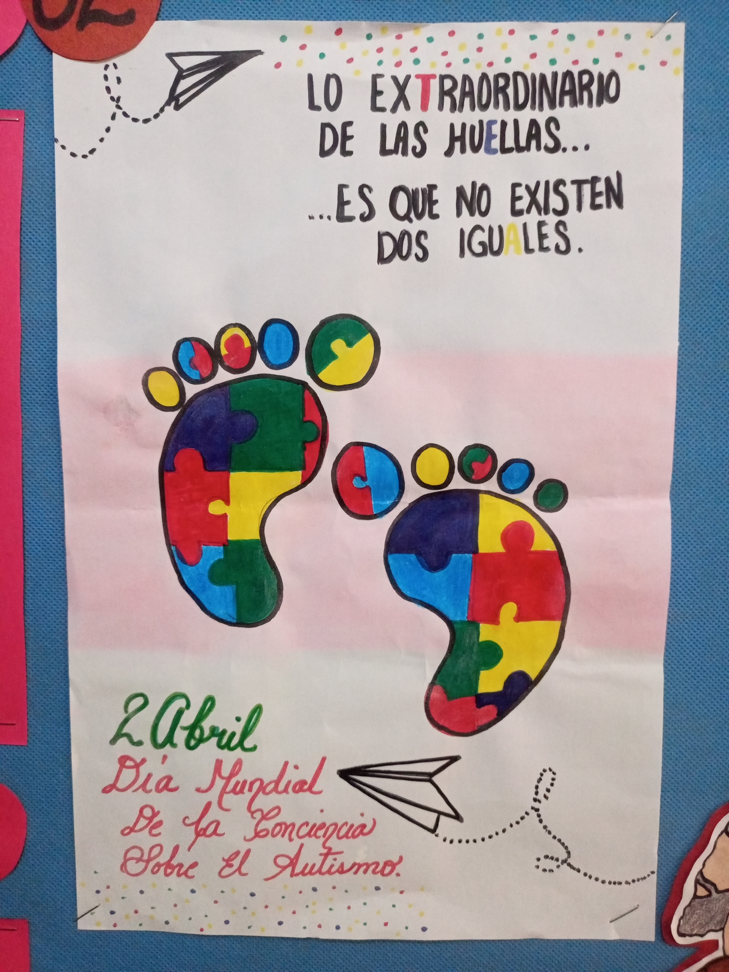 The image is a close-up photograph of a poster displayed on a blue textured surface, likely a bulletin board, in Santa Ana, Venezuela. The poster is white paper with colorful drawings and text. At the top, in large black letters, it reads "LO EXTRAORDINARIO DE LAS HUELLAS... ES QUE NO EXISTEN DOS IGUALES." Some of the letters are highlighted in different colors, such as "EXTRAORDINARIO" in red and blue, and "IGUALES" in yellow.
Below the text are two drawings of footprints, each made of puzzle pieces in red, yellow, blue, and green. The toes of the footprints are represented by colored circles. A black paper airplane is drawn to the right of the left footprint, with a dotted line trailing from it.
In the lower left corner of the poster, there is handwritten text in green and red. The green text reads "2 Abril," and below it, in red cursive, it says "Día Mundial De la Concienciación Sobre El Autismo." This translates to "April 2nd World Autism Awareness Day." Small colorful dots, similar to those at the top, are scattered below this text.
On the right side of the poster, a faint dotted line forms a shape resembling the letter "S," connecting to another paper airplane design. The edges of the poster are slightly curled, and there are some minor wrinkles in the paper. The blue background appears to be fabric or a similar textured material, common for classroom or community display boards. The lighting suggests an indoor setting.