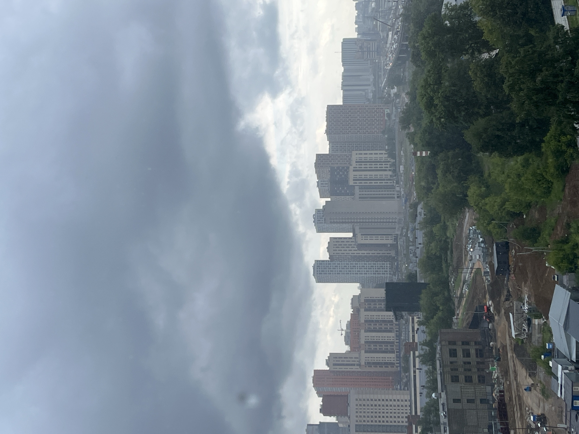 This media file depicts a cloudy, overcast day in Tarasovka, Russia. The scene is a developing urban area with numerous high-rise apartment buildings under construction or newly built. The buildings vary in height and architectural style, with some appearing to be completed and others in earlier stages of development. A prominent feature is a construction site with visible heavy machinery, dirt mounds, and partially built structures.
The sky is dominated by thick, gray clouds, suggesting a chance of rain or that it has recently rained. The lighting is diffused, creating a somewhat muted and somber atmosphere. Green trees are interspersed between the buildings and along the edge of the construction area, adding a natural element to the urban landscape. There are some indications of vehicular traffic, suggesting that this is an active area.
The perspective is from an elevated viewpoint, looking down and across the cityscape. The composition, with the buildings and construction stretching into the distance, conveys a sense of ongoing expansion and development. No people are clearly visible in the frame. The time of day is indeterminate due to the overcast conditions, but it appears to be daytime. No discernable text is present in the image.