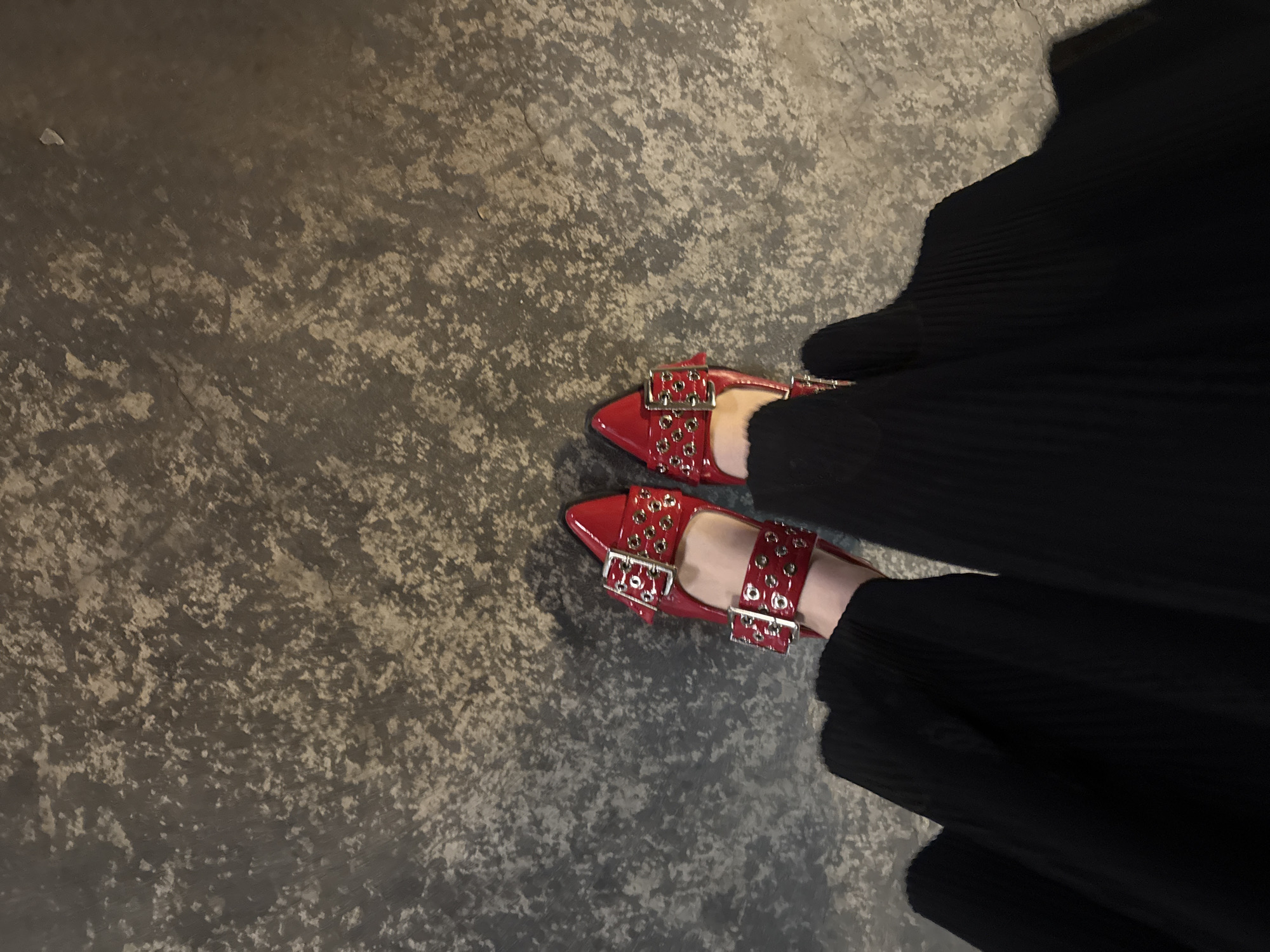 The image shows a person wearing red, patent leather shoes with silver buckles and a long, black pleated skirt. The shoes are on a textured gray surface. The location is unspecified, but the city is Mexico City, Mexico.