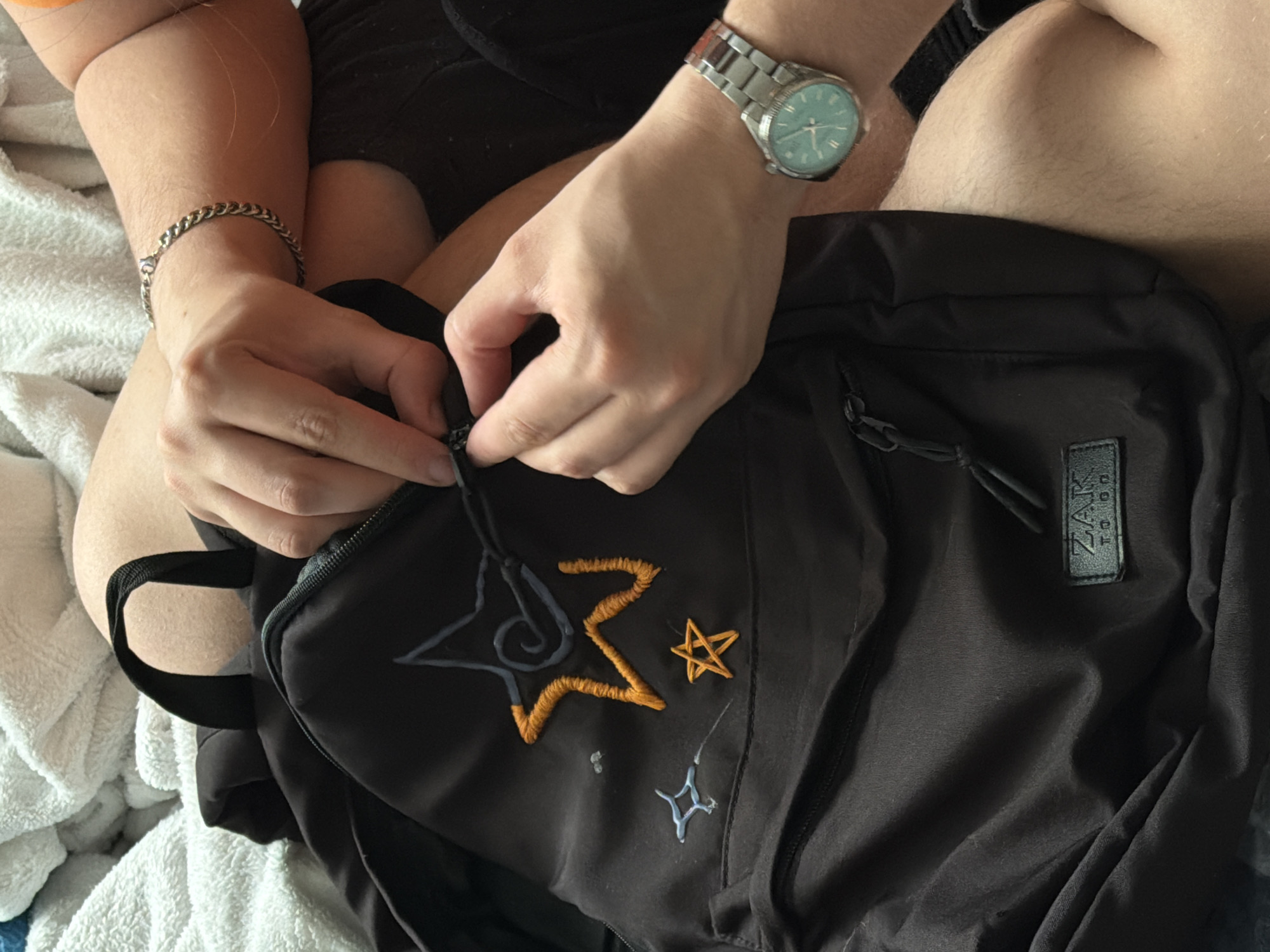 A person is shown from above, seated, with a black backpack on their lap. The person is wearing a silver bracelet on the left wrist and a silver watch with a light blue face on the right wrist. The person's hands are manipulating the zipper of the backpack. The backpack has a black label that reads "ZAK TO.DD." The backpack is decorated with embroidered designs, including stars and swirls in various colors like blue, yellow, and orange. The person is sitting on a white, textured surface, possibly a towel or blanket. The environment is indistinct. There is no indication of the location being Perm, Russia.