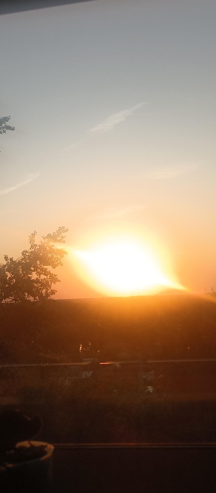 The image depicts a sunrise or sunset in an outdoor setting. The sun is positioned near the horizon, emitting a bright, diffused light. Silhouetted against the sun are trees and a low-lying hill or embankment. The sky transitions from a light blue to orange near the horizon. A faint contrail is visible in the upper part of the sky. The foreground is dark, with a partial view of a vehicle interior, including what appears to be a dashboard and a container.