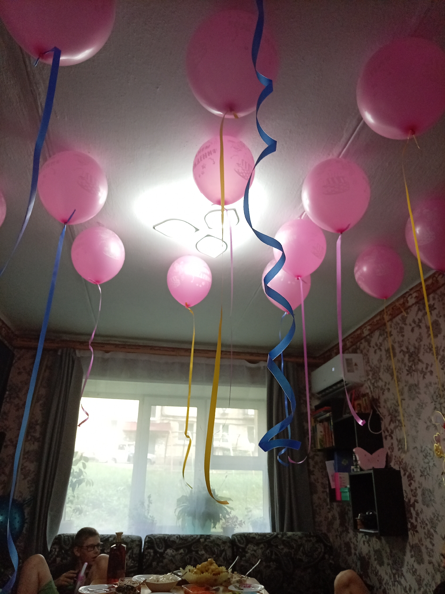 This image depicts a ceiling decorated with numerous pink balloons and colorful ribbons, creating a festive atmosphere. The balloons are of various sizes and some have imprinted designs. In the background, a window offers a view of a residential area, suggesting an indoor setting within a city. A shelf unit filled with books and decorative items is visible on the right side of the frame. The overall impression is one of a celebration or party.