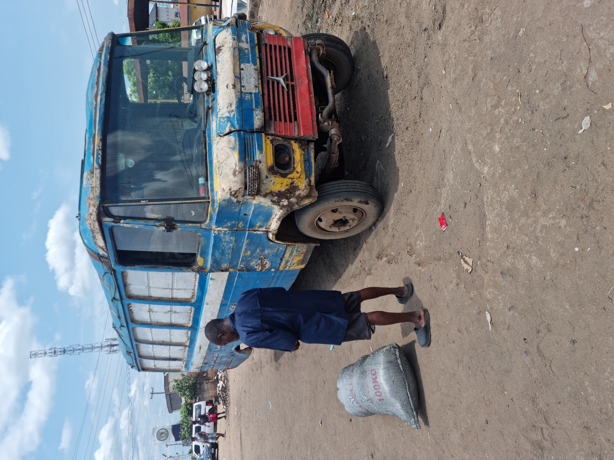 This outdoor image depicts a dilapidated, heavily rusted blue and yellow bus or truck parked on a dirt road or unpaved lot under a bright, partly cloudy daytime sky. The vehicle shows significant wear, with peeling paint and exposed parts, and features a red front grill with a star-like emblem and three auxiliary lights mounted above its dirty windshield. Its side windows appear covered or opaque white.
A man with dark skin stands beside the bus, facing away from the camera, wearing a dark blue long-sleeved shirt, dark shorts, and dark slip-on shoes. Near his feet, a large grey woven sack lies on the ground, clearly marked with "400KG." In the background, power lines, a tall metal structure, and parts of buildings are visible, suggesting an urban or semi-urban environment. The scene implies a moment of daily activity, possibly related to commerce or transport, within a working area, consistent with the context of Onitsha, Nigeria. No strong emotions are discernible.