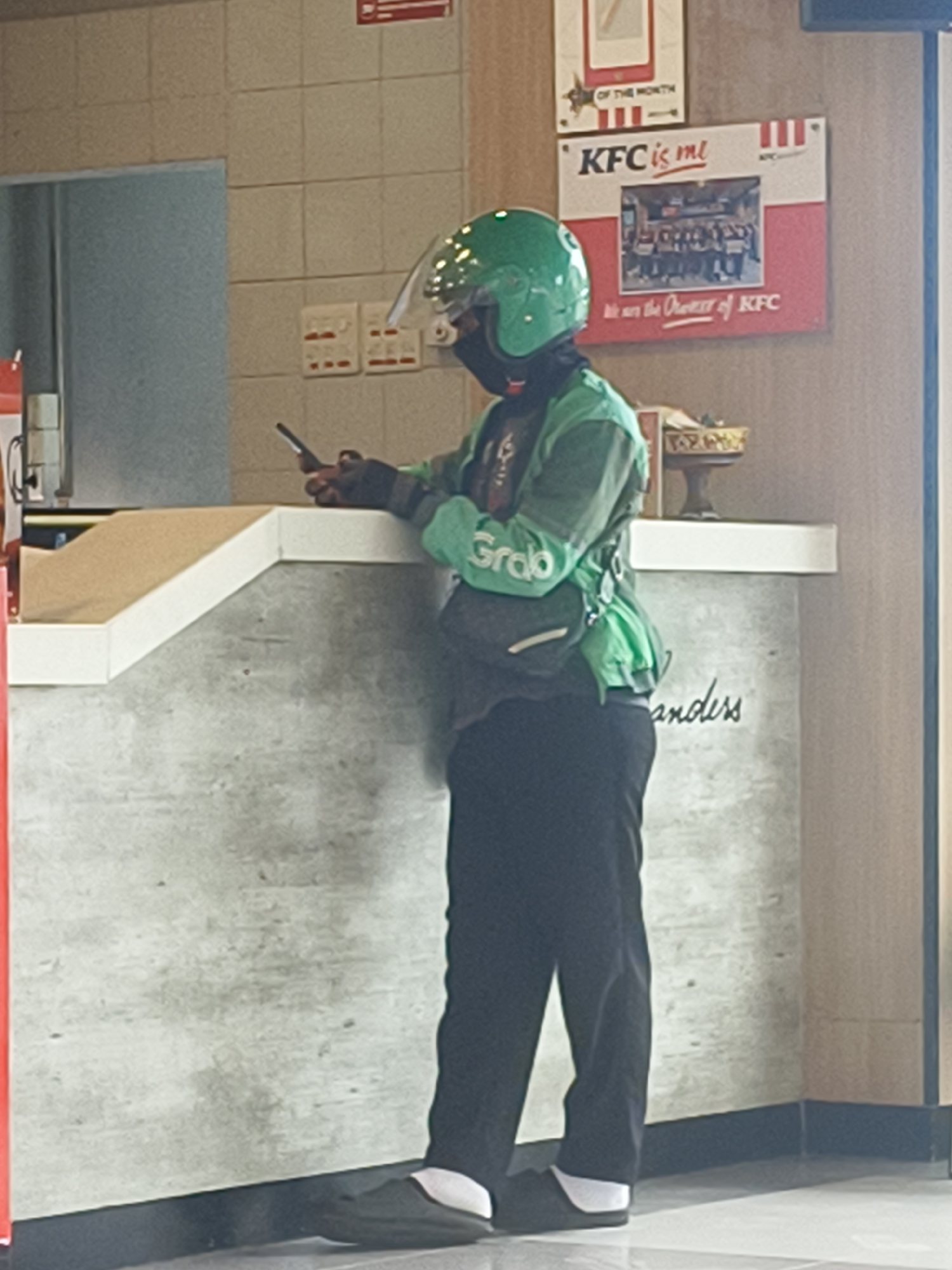 A person wearing a green helmet and a green jacket with "Grab" logo is standing at a counter inside a KFC restaurant. The person is holding a mobile phone in their right hand and looking at it. The counter is made of concrete and white material. There are posters on the wall behind the person, one of them saying "KFC is me". The floor is tiled. The scene is indoors.