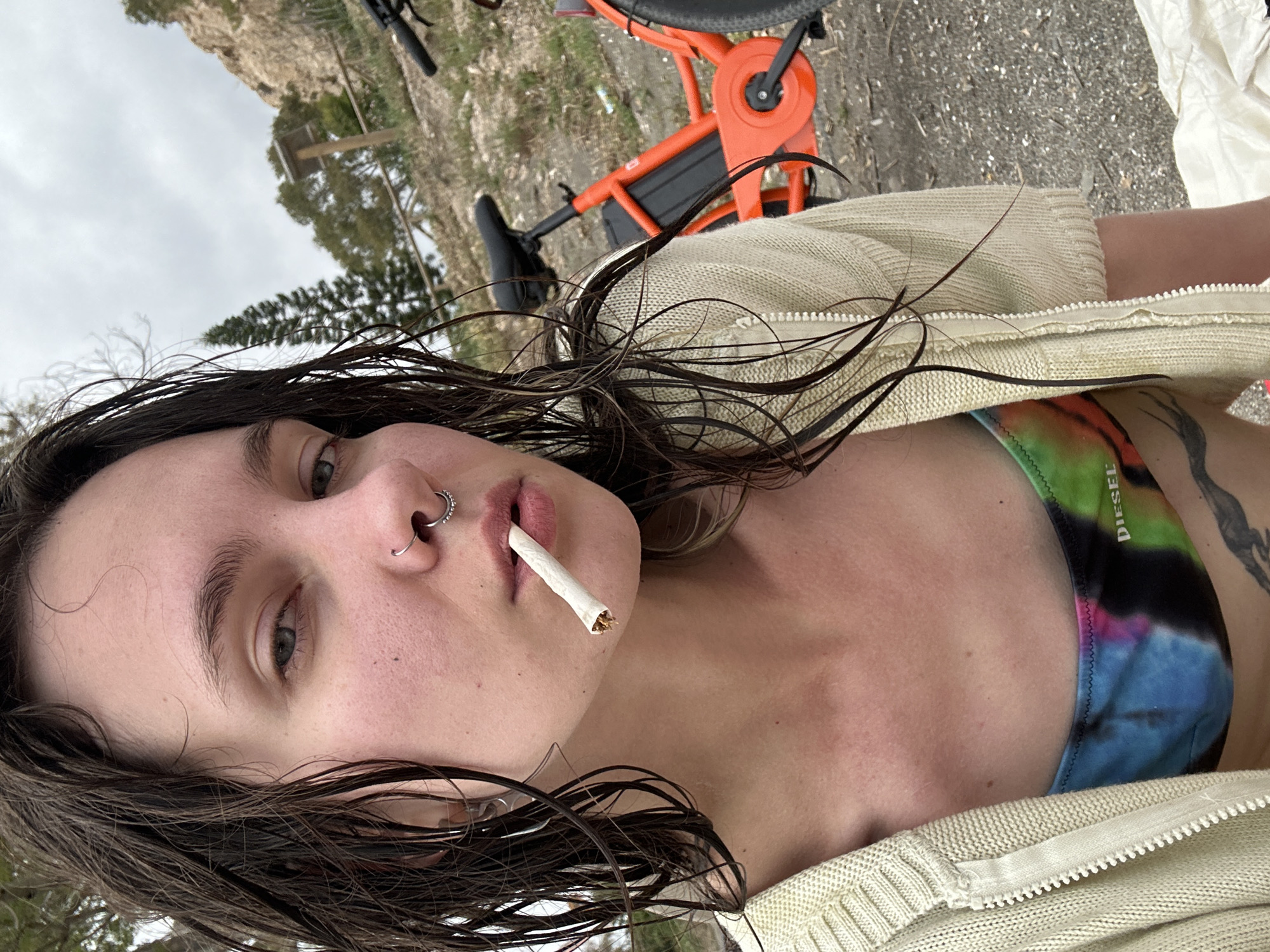 An eye-level portrait shows a person with wet, dark hair and light eyes looking directly at the camera. They have a neutral expression, with a hand-rolled cigarette or joint held between their lips. The person features both a septum piercing (circular barbell) and a ring in their left nostril. They are wearing a light-colored, possibly cream or beige, knit cardigan or hoodie, partially unzipped, revealing a colorful tie-dye bikini top or bralette underneath. The text "DIESEL" is visible on the top. A black, abstract, or tribal-style tattoo is visible on their left upper arm/shoulder.
The background is an outdoor setting, likely a rocky or gravelly path, with sparse vegetation and trees under an overcast sky. Behind the person, an orange electric bicycle with black components (seat, battery pack, handlebars) is partially visible. The wet hair suggests recent water activity, possibly at a beach or near water, consistent with a coastal location like Málaga, Spain. The overall mood is casual and relaxed.