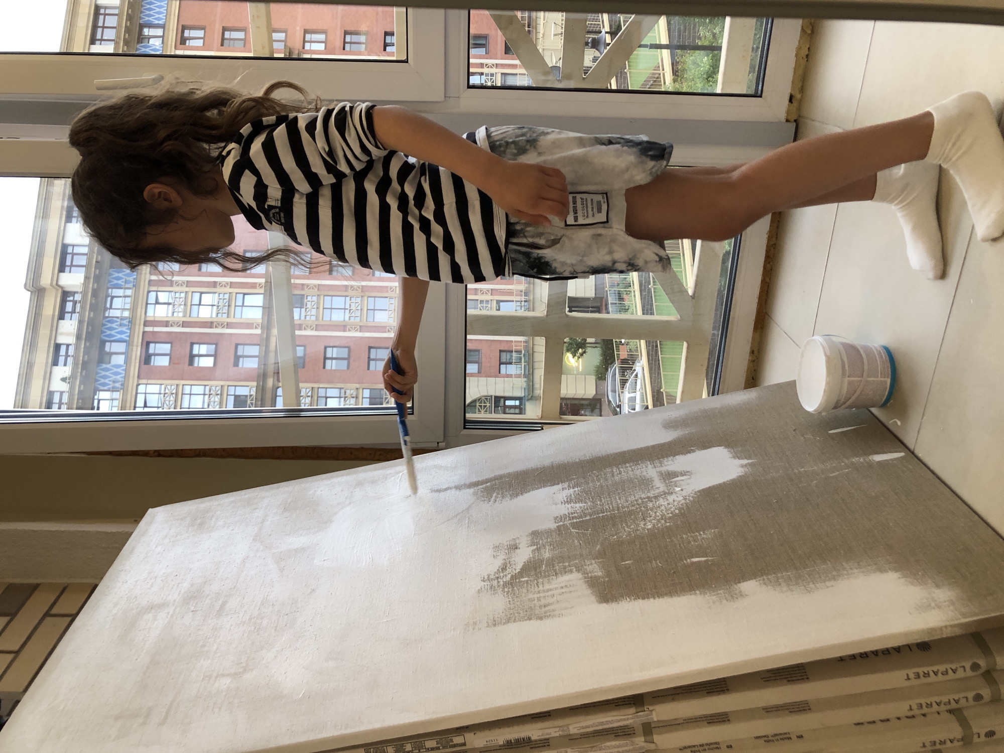 A young girl with curly brown hair tied in a ponytail, wearing a black and white striped long-sleeve shirt and tie-dye shorts, stands indoors next to a large canvas. She is in the process of painting the canvas with a roller, applying white paint. To the left of the canvas are stacked packages, possibly building materials, with "LAPARET" visible on them. A small white bucket with a blue lid sits on the floor near the canvas. The girl is standing on a tiled floor. Behind her is a large window overlooking a city street with brick buildings and trees. The scene appears to be an interior space with natural light streaming in from the window. The architecture visible through the window suggests a European city, possibly Saint Petersburg, Russia, given the style of the buildings.