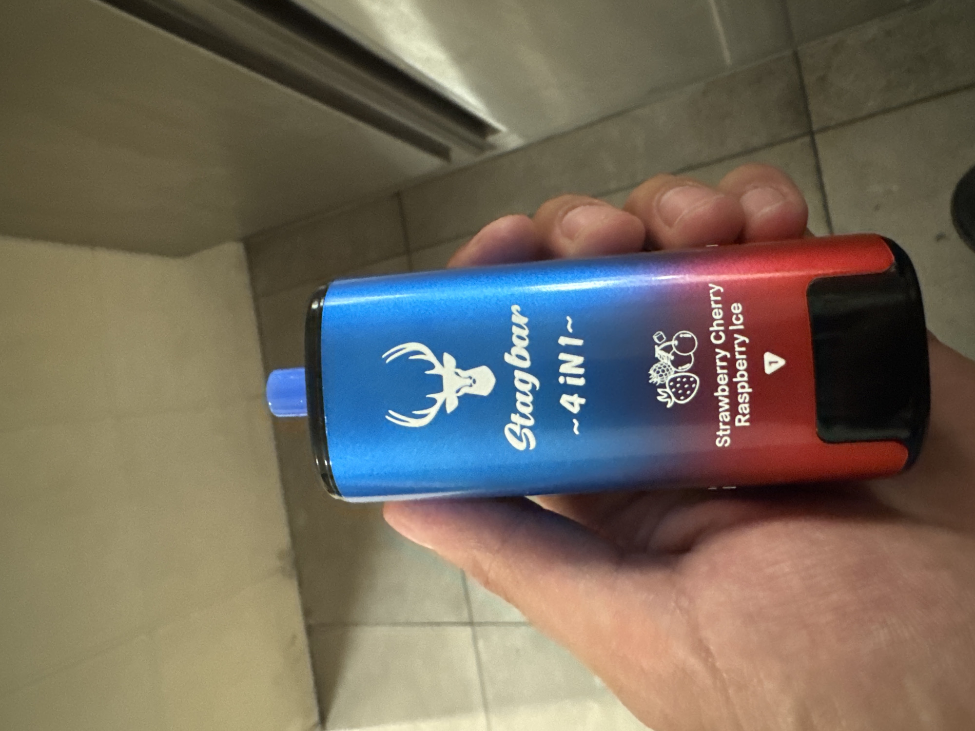 A person is holding a "Stagbar - 4 in 1" disposable vape, with a flavor description of "Strawberry Cherry Raspberry Ice." The vape is blue on one end and fades into red on the other. The background consists of beige wall panels on the left and tiled flooring on the right. The photo appears to be taken indoors. It is not possible to determine if this is Sofia, Bulgaria, or any other city based on the provided image.