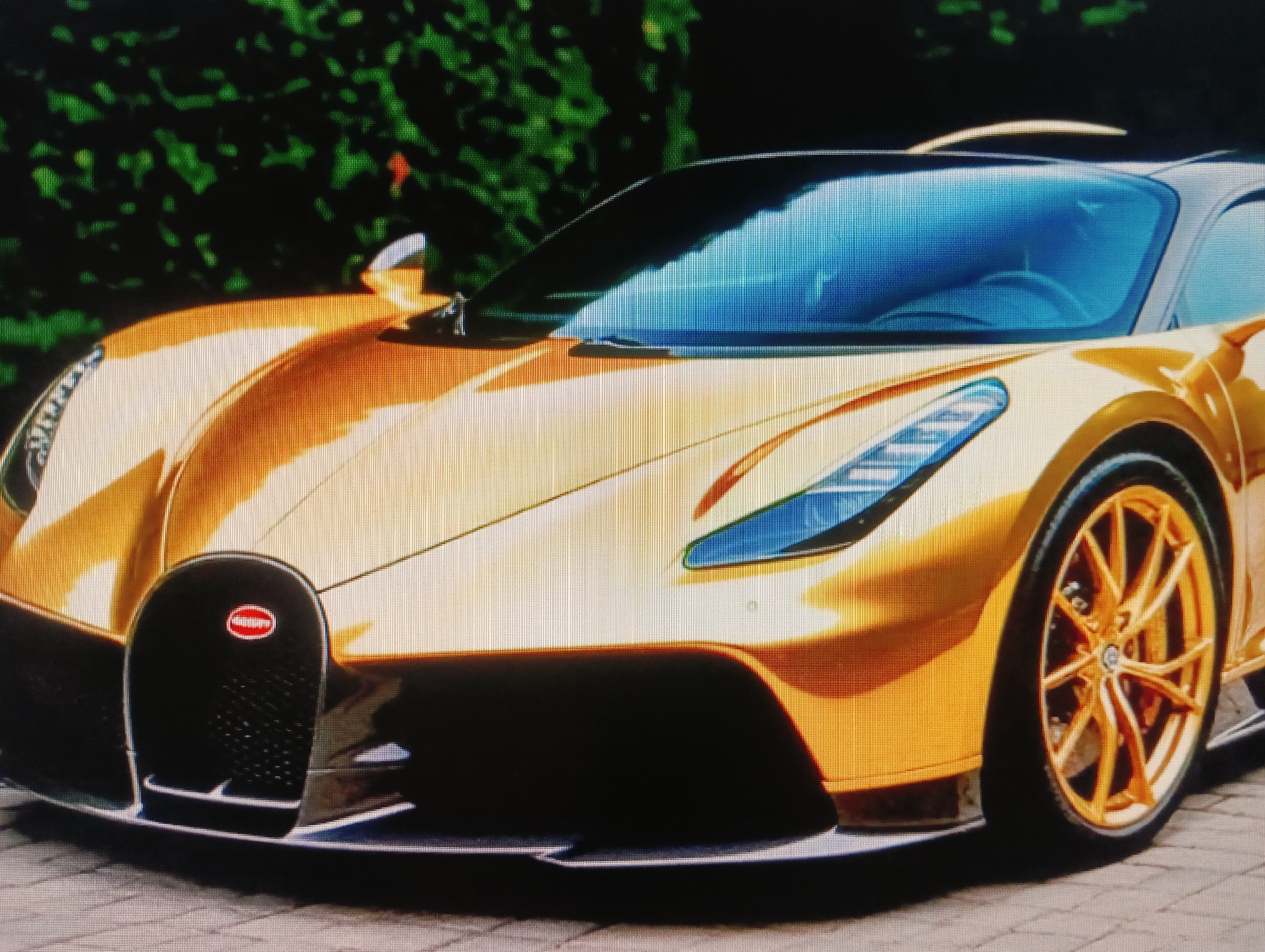 The image shows a close-up, eye-level, medium shot of a golden Bugatti Chiron Super Sport 300+ parked on a paved surface. The car's front grille, headlights, windshield, and front passenger-side wheel are visible. The car is positioned outdoors, with dark green foliage in the background. The lighting suggests it is daytime, and the car's metallic gold finish reflects the surroundings. No people are present in the image. The location is not specified as Sluck, Belarus.