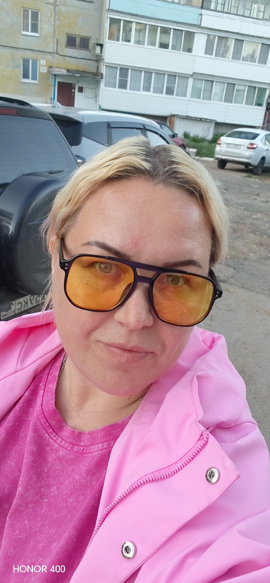 A close-up outdoor portrait captures an adult woman with blonde hair and dark roots, wearing prominent yellow-tinted sunglasses with dark frames. She is dressed in a bright pink t-shirt layered under a lighter pink, shiny jacket with a zipper and snap buttons. A silver chain is visible around her neck. Her expression is neutral, looking directly at the camera.
The background reveals a typical Russian urban or suburban setting, confirming the provided location context of Noviy, Russia. Behind her, several cars are parked, including a dark SUV or car to her left with a partially visible Cyrillic license plate (appearing to start with "ОЯРС" or "ОЯКС"). Further back, a silver sedan is visible. Beyond the cars, multi-story apartment buildings, characteristic of Russian architecture, line the street. One building is a weathered yellowish-grey, while another appears white with numerous windows and enclosed balconies. The ground consists of asphalt or gravel, with some greenery in the distance. The lighting suggests it is daytime, possibly late afternoon, with diffused light.
Visible text includes a "HONOR 400" watermark in the bottom left corner of the image.