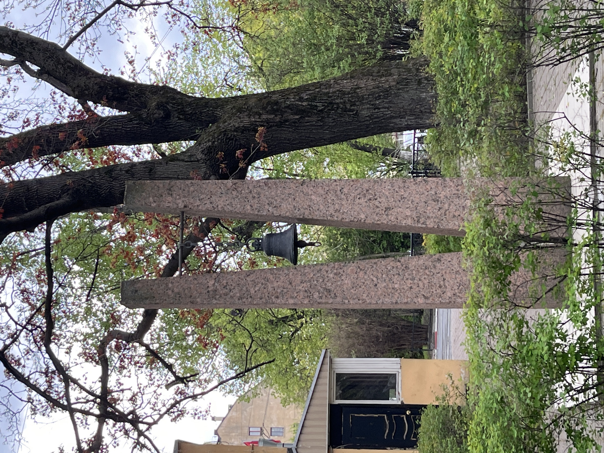 The image depicts the ringing bell monument in Saint Petersburg, Russia. The monument comprises two light brown, granite pillars slanted towards each other with a bell suspended between them. A large tree with budding leaves is behind the pillars. Below the pillars, parts of buildings are visible, including a small structure with a black door and a window, and portions of other buildings in the background. Green foliage is also present, partially obscuring the view. The sky is visible through the tree branches and appears overcast.
