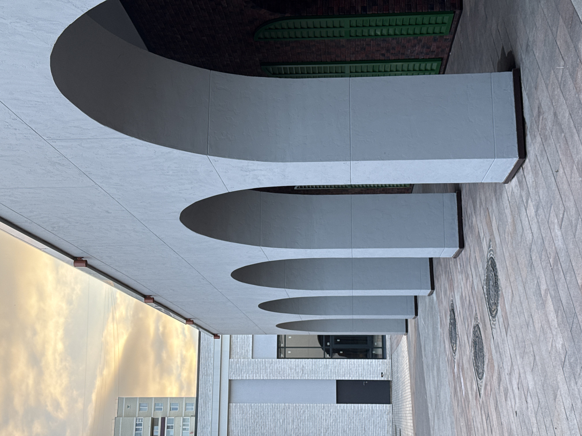 The image depicts an architectural detail of a building with a series of arched openings. The arches are rendered in a light-colored, textured material, creating a rhythmic pattern across the facade. The building appears to be modern, with clean lines and a contemporary aesthetic.
The scene is set outdoors, with a paved area in the foreground, composed of interlocking stones in varying shades of brown and gray. Three circular, decorative manhole covers are embedded in the paving stones, adding a subtle design element to the ground.
Above the arches, the building's upper structure is visible, featuring dark brickwork and green architectural elements that suggest a rooftop or canopy. The contrast between the light arches and the darker upper section creates a visual depth.
To the left of the frame, a wide expanse of sky dominates the background. The sky is filled with dramatic, golden clouds, indicative of either sunrise or sunset. The warm hues of the sky suggest a pleasant time of day, likely during the golden hour. Partially visible at the bottom left is a modern, multi-story building with a light-colored facade and numerous windows, suggesting an urban or suburban environment.
The image does not contain any people or overt signs of activity. The focus is purely on the architectural design and the interplay of light and shadow on the building and its surroundings. The overall mood is one of calm and visual interest, highlighting the geometric patterns and the atmospheric lighting. There is no visible text in the image. The location context of Izhevsk, Russia, is noted but not visually evident within the image itself.