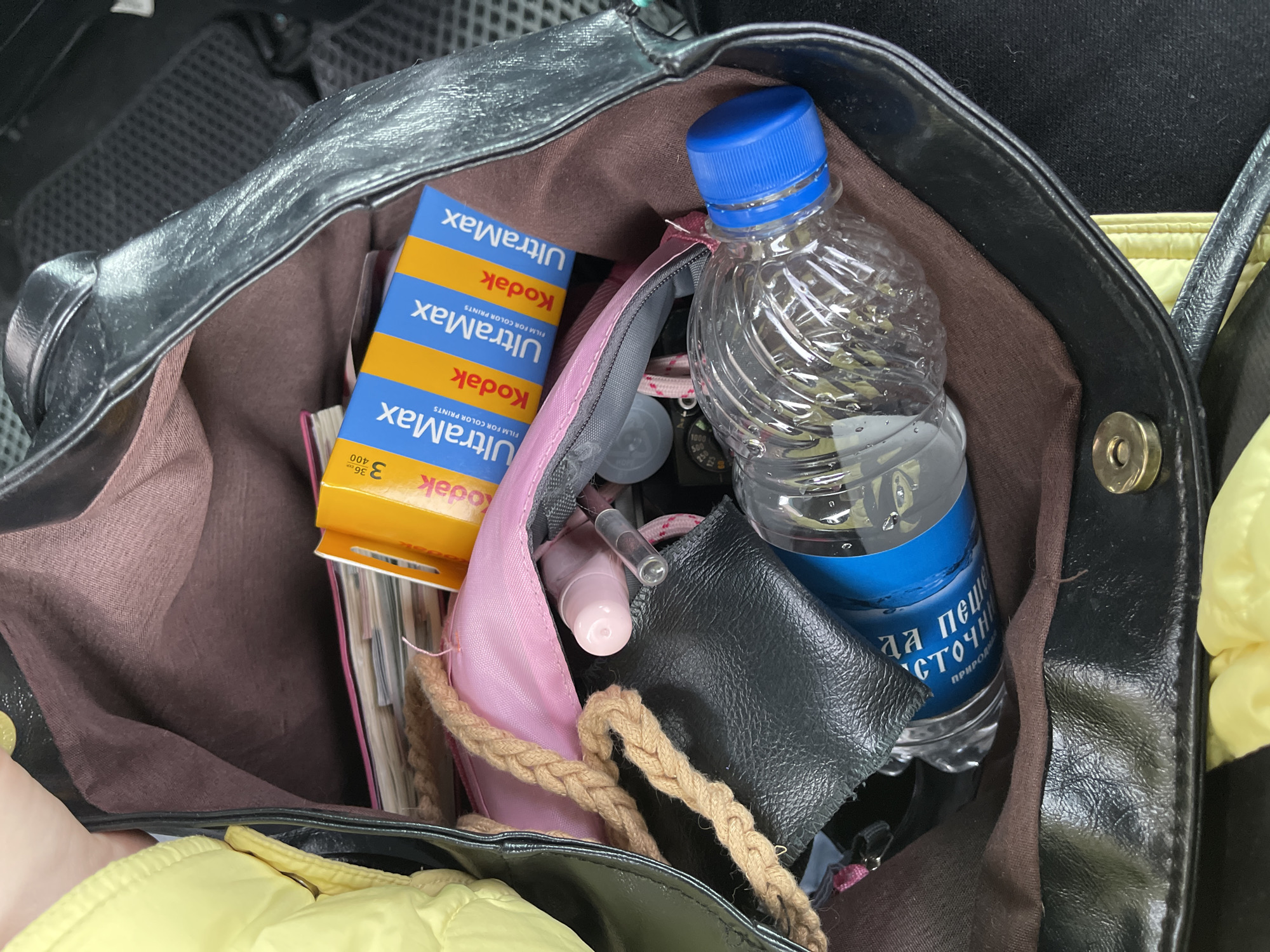 The image shows a black leather handbag filled with various items. Inside, there are three boxes of Kodak Ultramax 400 film, a pink and gray makeup bag, a water bottle with a blue cap, and a small black wallet. A notebook is also visible near the film boxes. The water bottle has Cyrillic text on it, which reads "да пеще источни" and "ПРИРОДНАЯ". The location appears to be inside a car, as a car mat is visible in the background, and the surrounding elements suggest an interior space. The image does not contain enough details to determine its specific location within Moscow, Russia.