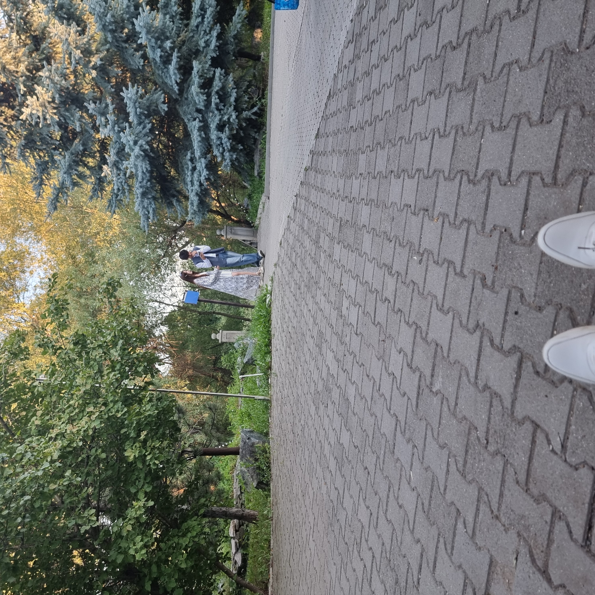 In the city of Aktobe, Kazakhstan, a couple walks along a paved pathway in a park or garden area. The pathway is constructed of interlocking gray concrete pavers. The woman on the left wears a long, patterned white dress and appears to be looking to her left. The man on the right, wearing a blue vest over a white shirt and jeans, is looking down and holding something to his face, possibly a phone. Behind them, a blue sign is mounted on a pole. The environment includes mature trees with green and yellow foliage, and a large blue spruce tree on the right. The perspective is low, suggesting the photo was taken from a seated or crouching position, with the viewer's white shoes visible in the foreground.