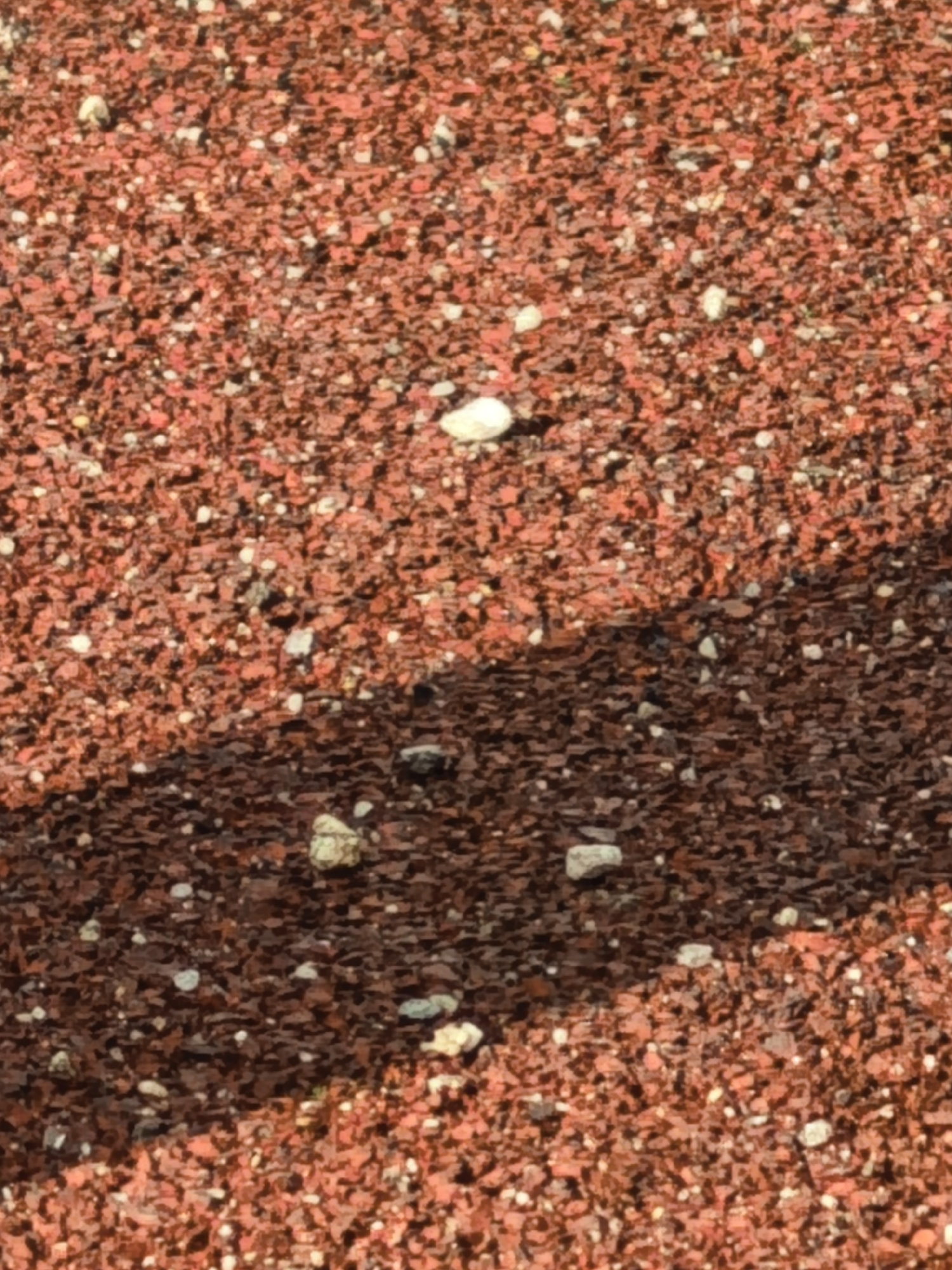 The image displays a close-up, aerial view of reddish-brown gravel with scattered white pebbles. A diagonal shadow, likely cast by an unseen object, divides the frame. The texture is granular and uneven, suggesting a natural or man-made ground cover. No people, animals, or discernible structures are present. The overall impression is of an outdoor surface under bright lighting conditions, indicated by the sharp shadow. The location is unknown, within a city of unknown name.