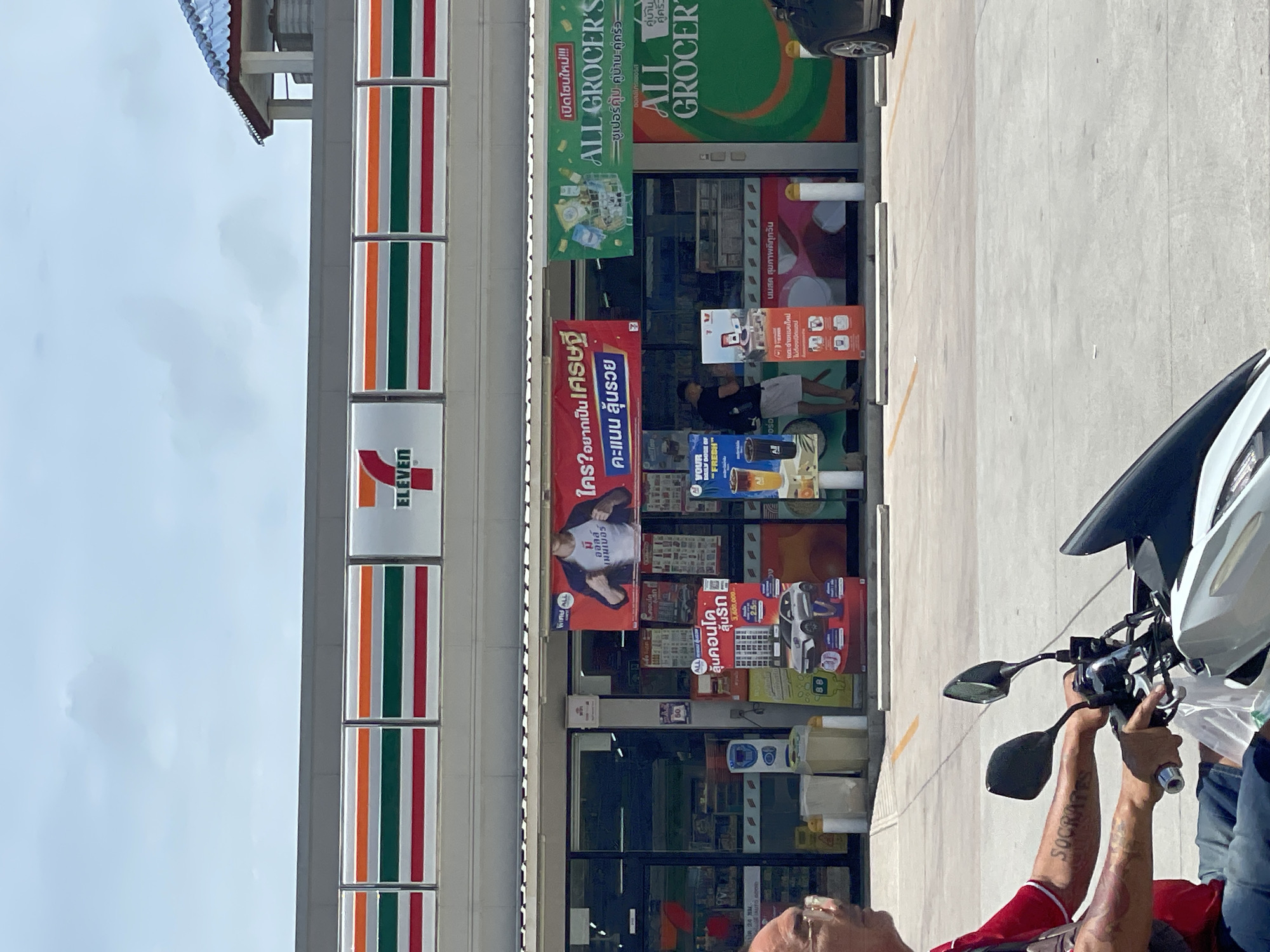 The image shows a 7-Eleven convenience store located in Pattaya City, Thailand, on a bright, overcast day. The store features the iconic red and white 7-Eleven logo prominently displayed on its facade, along with colorful promotional banners. One banner, in Thai, advertises a promotion for "ALL GROCER'S." Inside the store, various products are visible on shelves through the large glass windows. Outside the store, there are several advertising displays, including one featuring a car and another with beverages. A person is seen standing in front of the store, appearing to be interacting with a display. In the foreground, a person on a motorcycle is captured in motion, with their arm bearing a tattoo that reads "SOCRATES." The ground is paved, and parking lines are visible. The overall environment suggests a commercial area in Pattaya City.